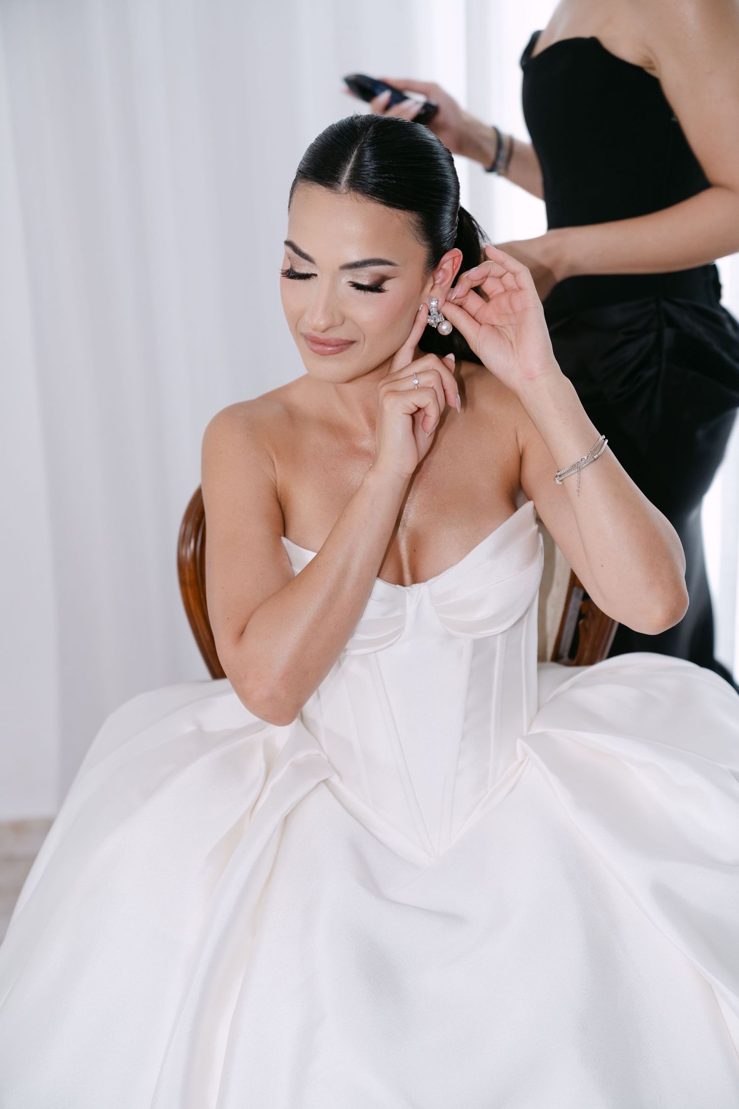 Bride in white corset gown having earrings put on by bridesmaid before wedding.