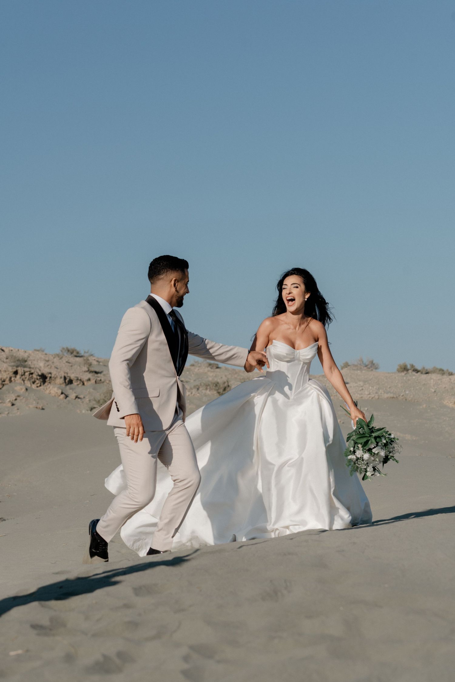 Laughing bride in white ballgown runs with groom across sand dunes under a bright blue sky.