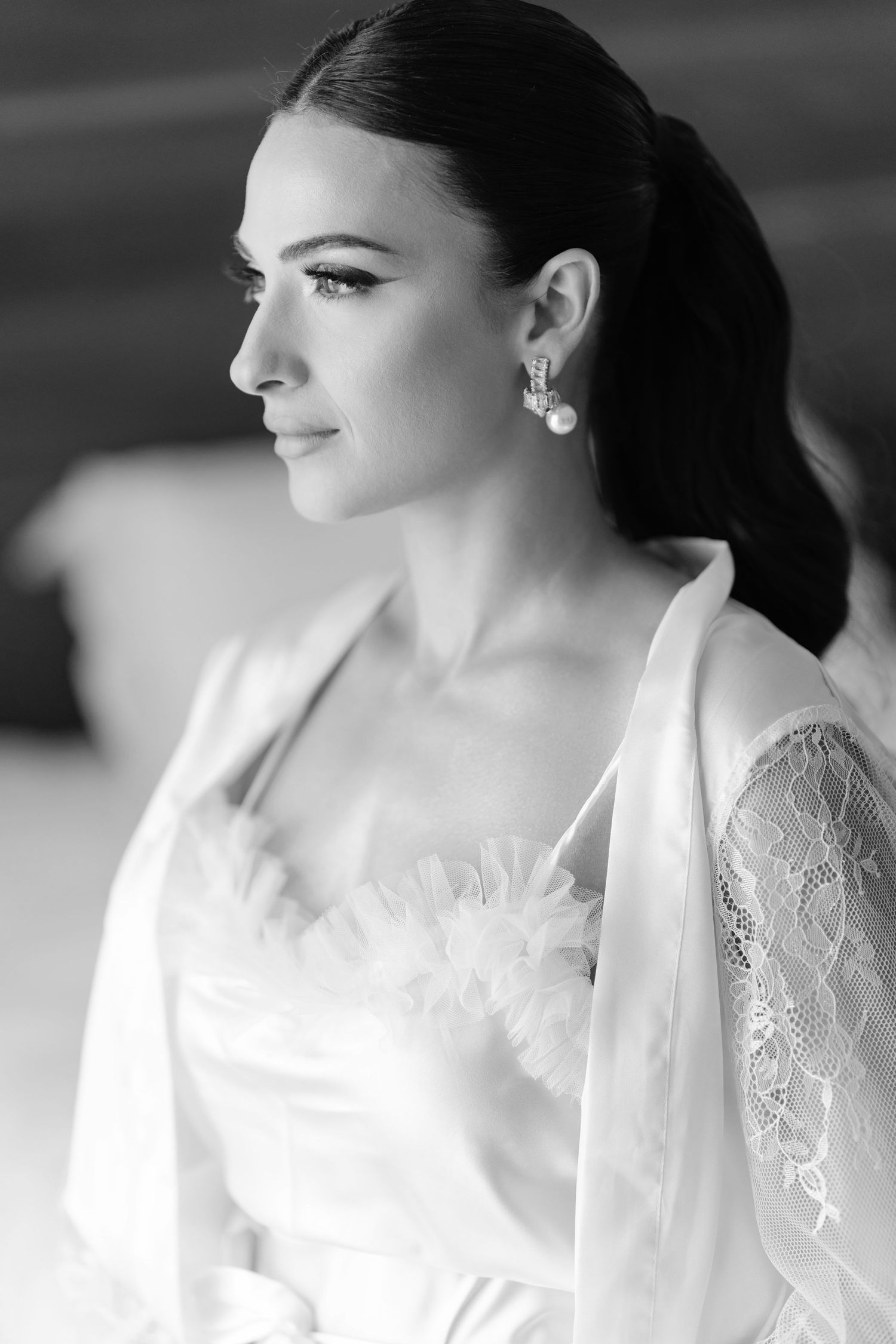 Elegant bride in white robe with lace sleeves and pearl earrings in black and white portrait.