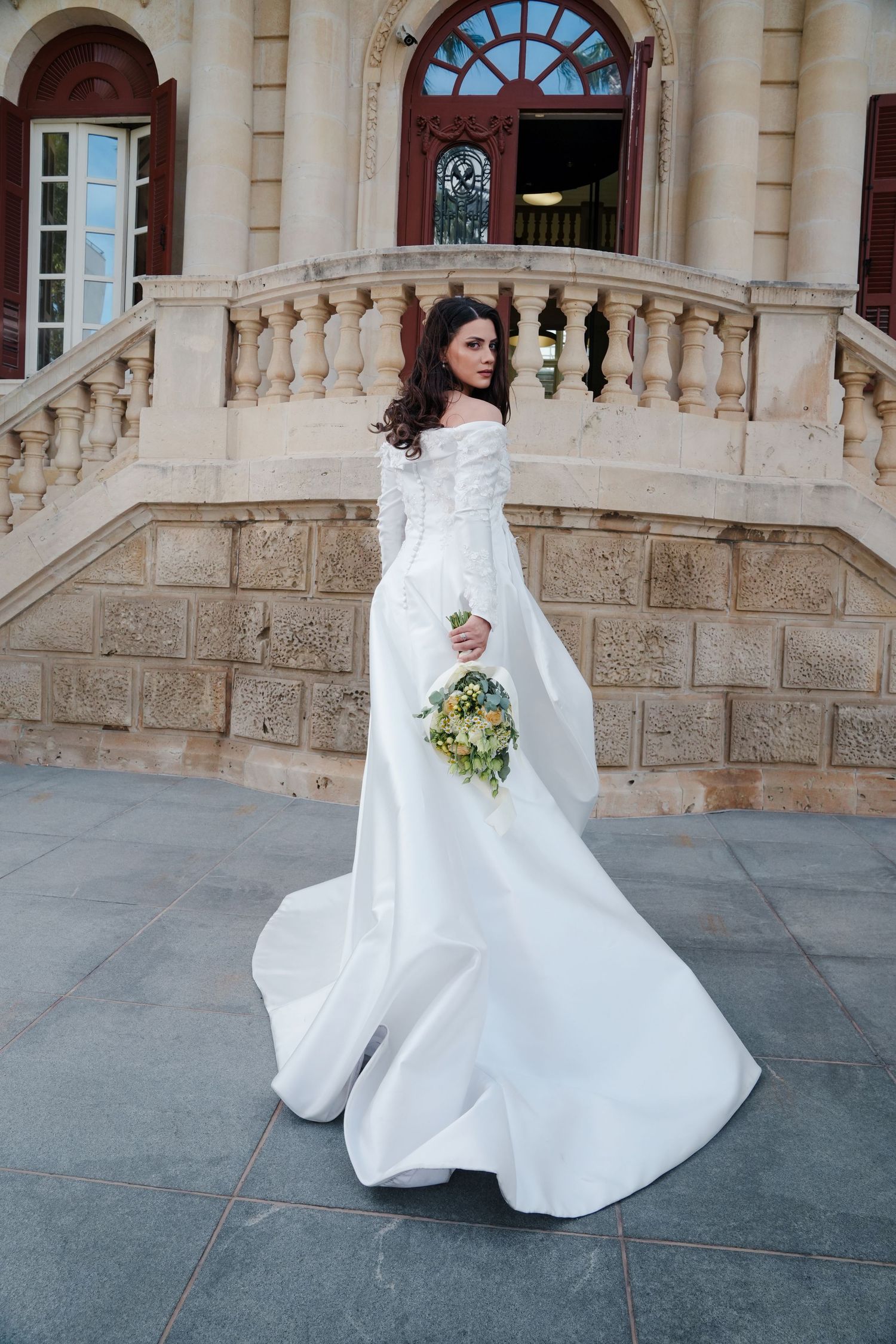 Elegant bride in a long-sleeve off-shoulder white wedding gown holding a bouquet on ornate stone steps.