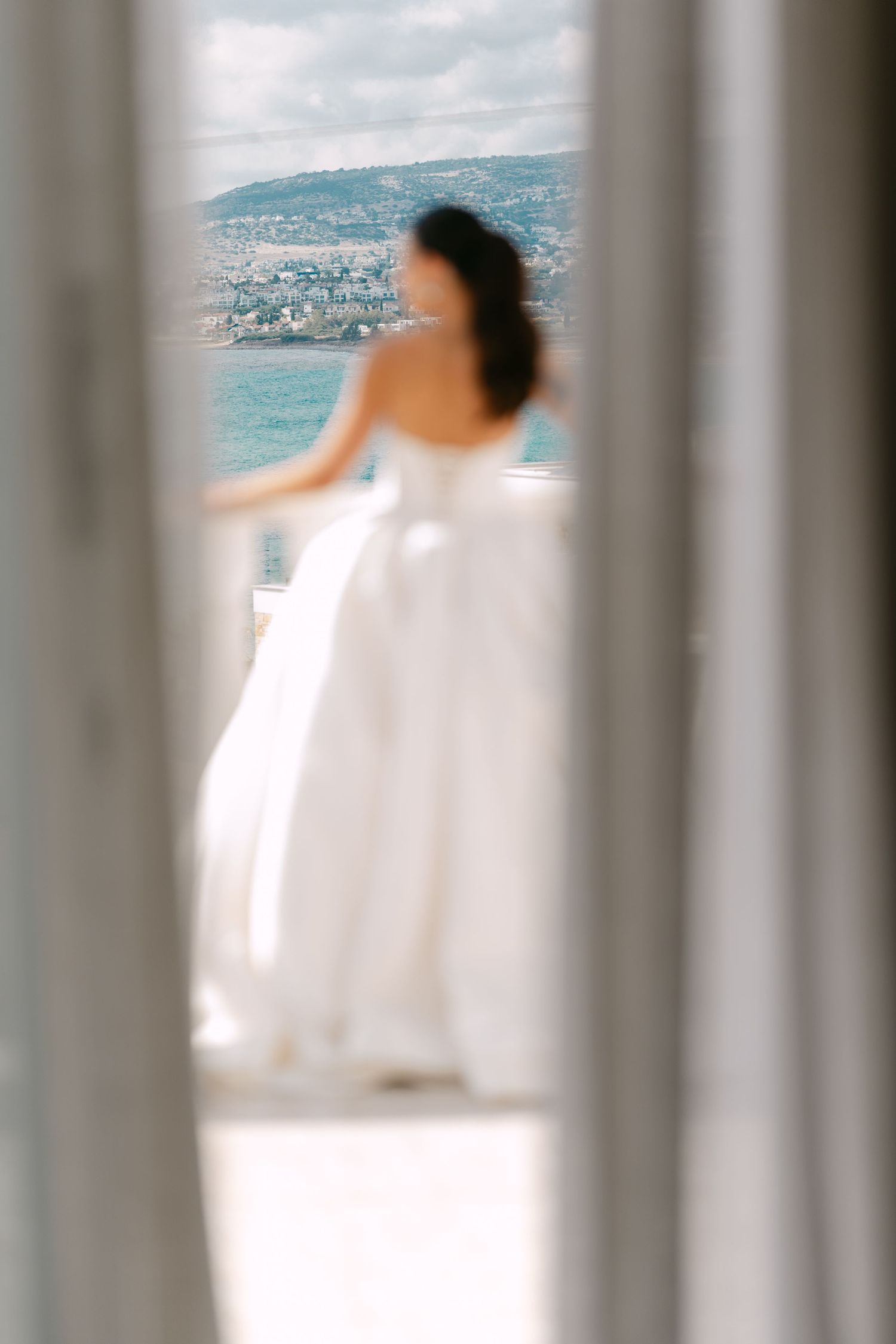 Bride in white wedding gown standing on balcony overlooking coastal cityscape.
