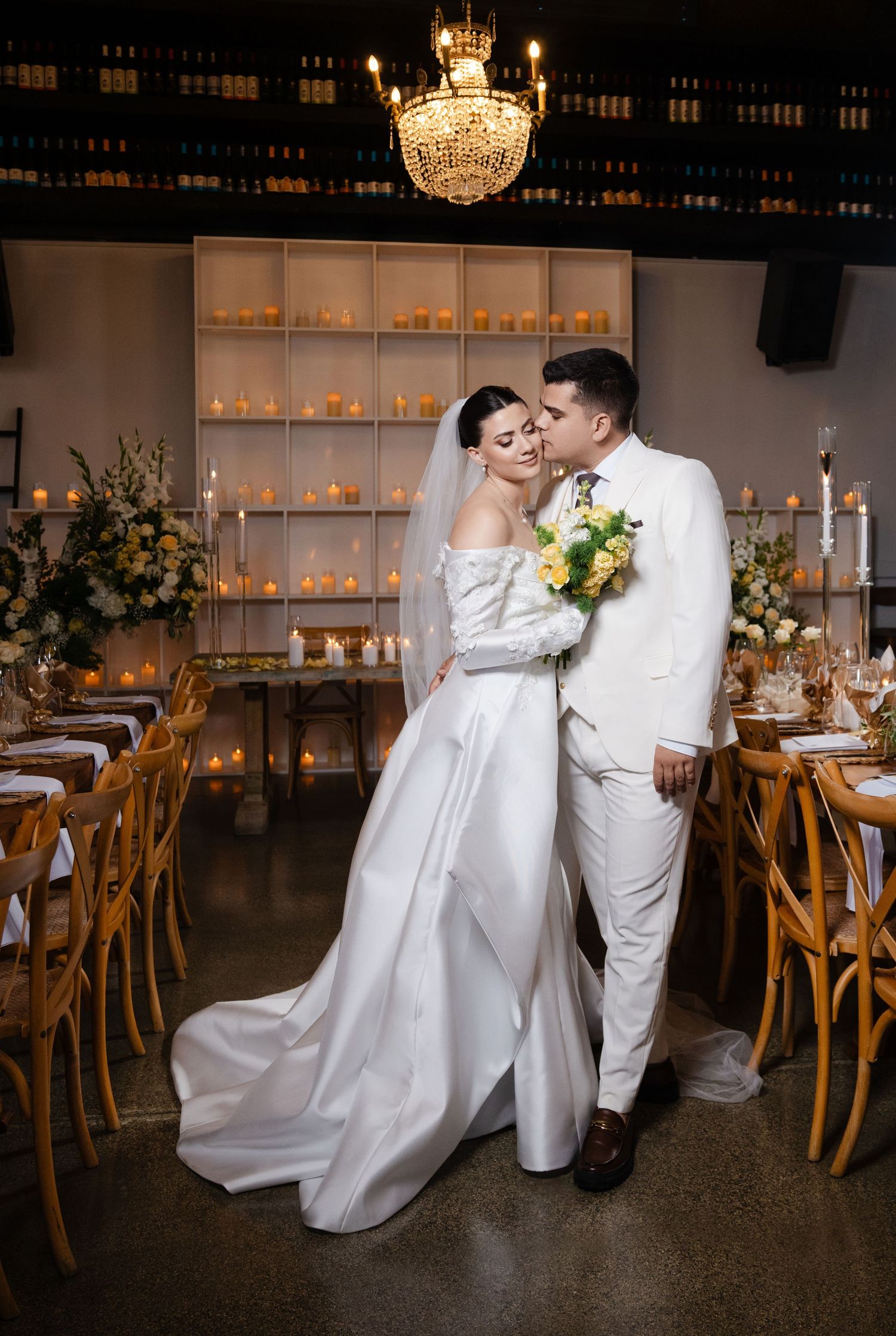 Bride and groom in white attire share a romantic moment holding a bouquet in a candlelit reception venue.