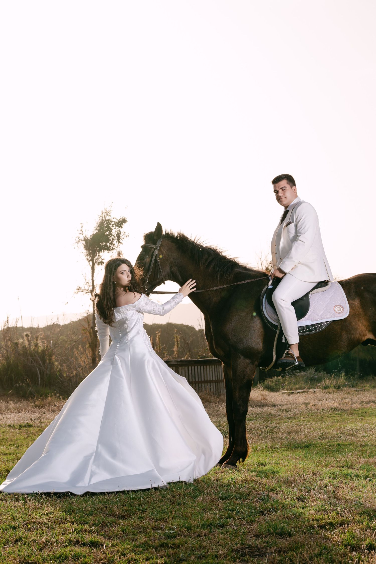 Bride in white ball gown touches dark horse as groom in white suit rides in golden sunset light.