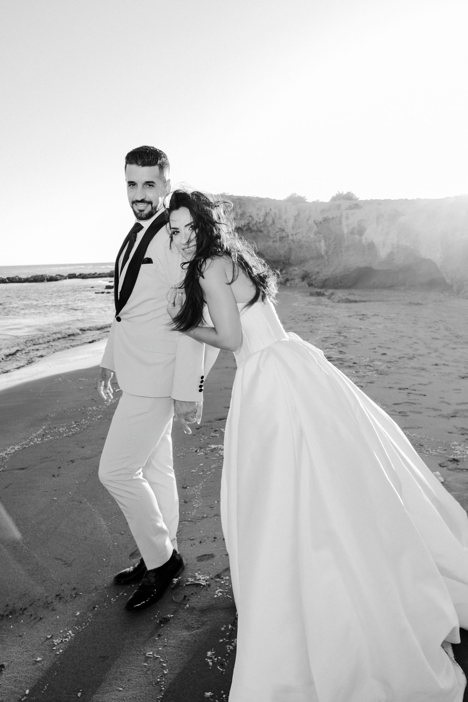 Black and white photo of smiling bride and groom walking playfully together on sandy beach shoreline.