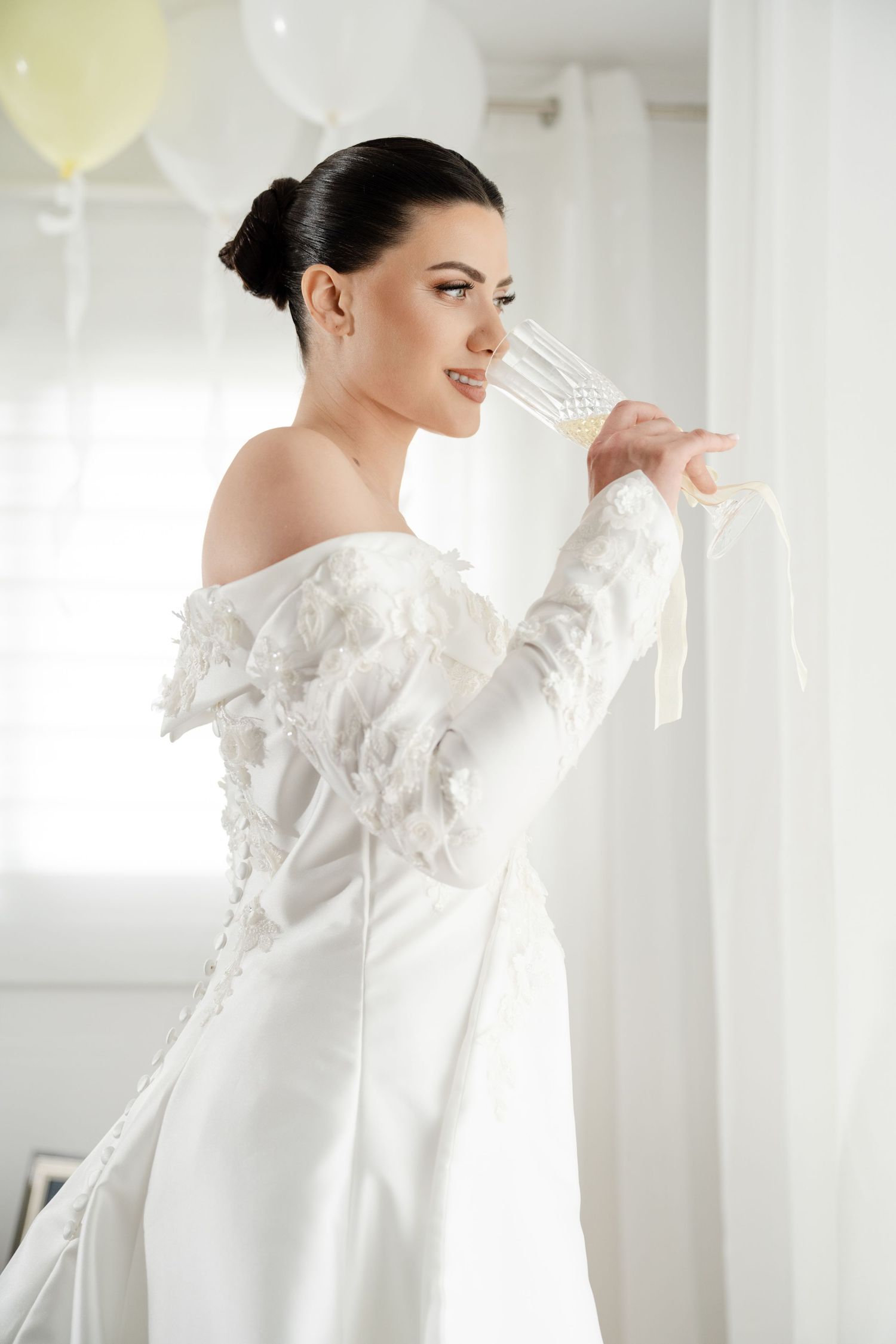 Beautiful bride in off-shoulder lace wedding gown holding champagne glass by window.