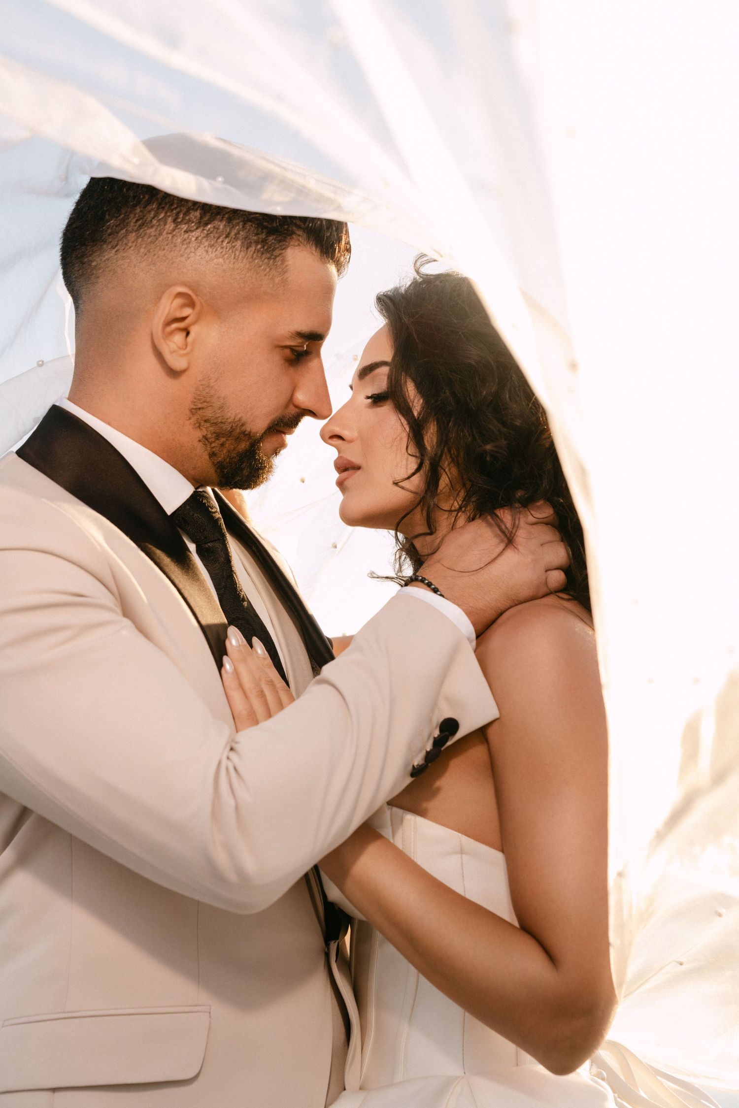 Bride and groom share an intimate romantic moment under flowing veil in warm golden sunlight.