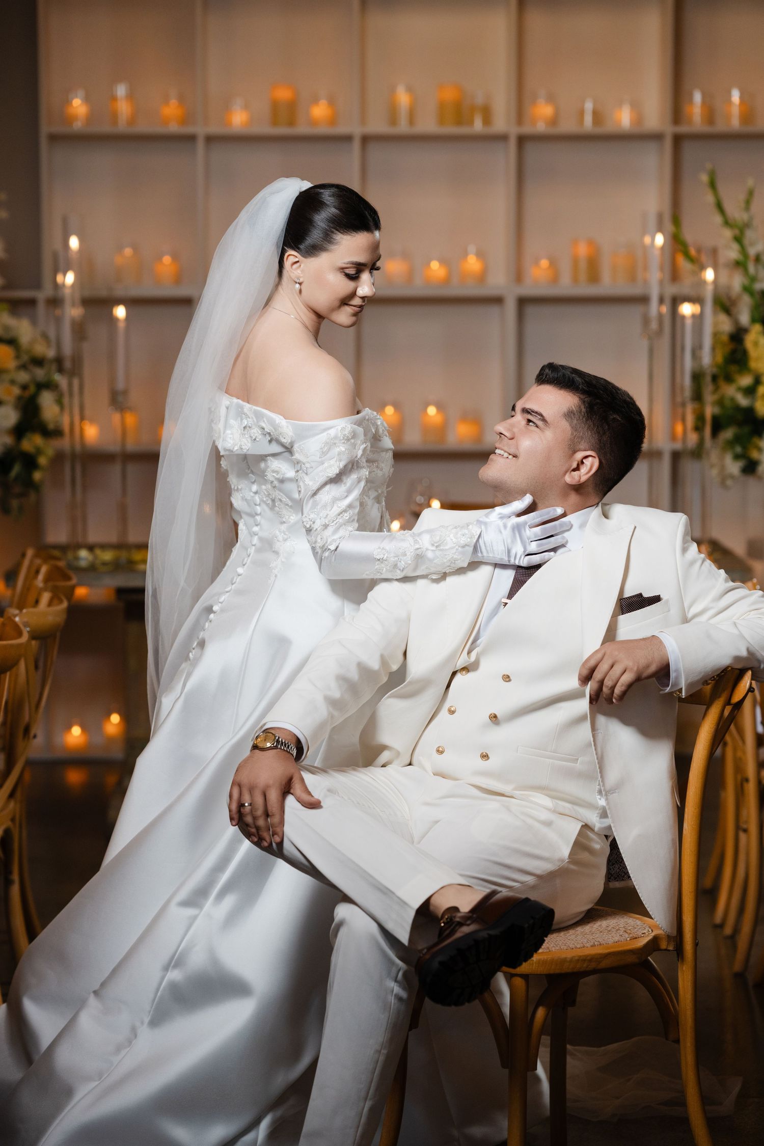 Bride in lace-sleeved gown smiles at seated groom in white suit surrounded by glowing candles at reception.