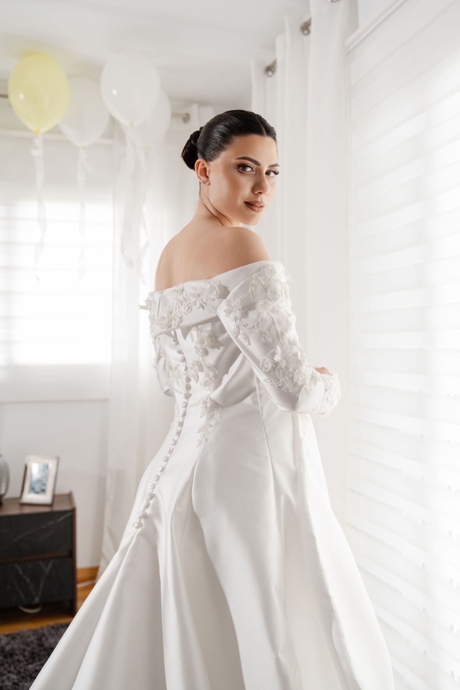 Elegant bride in off-shoulder lace wedding gown with button back posing by window.
