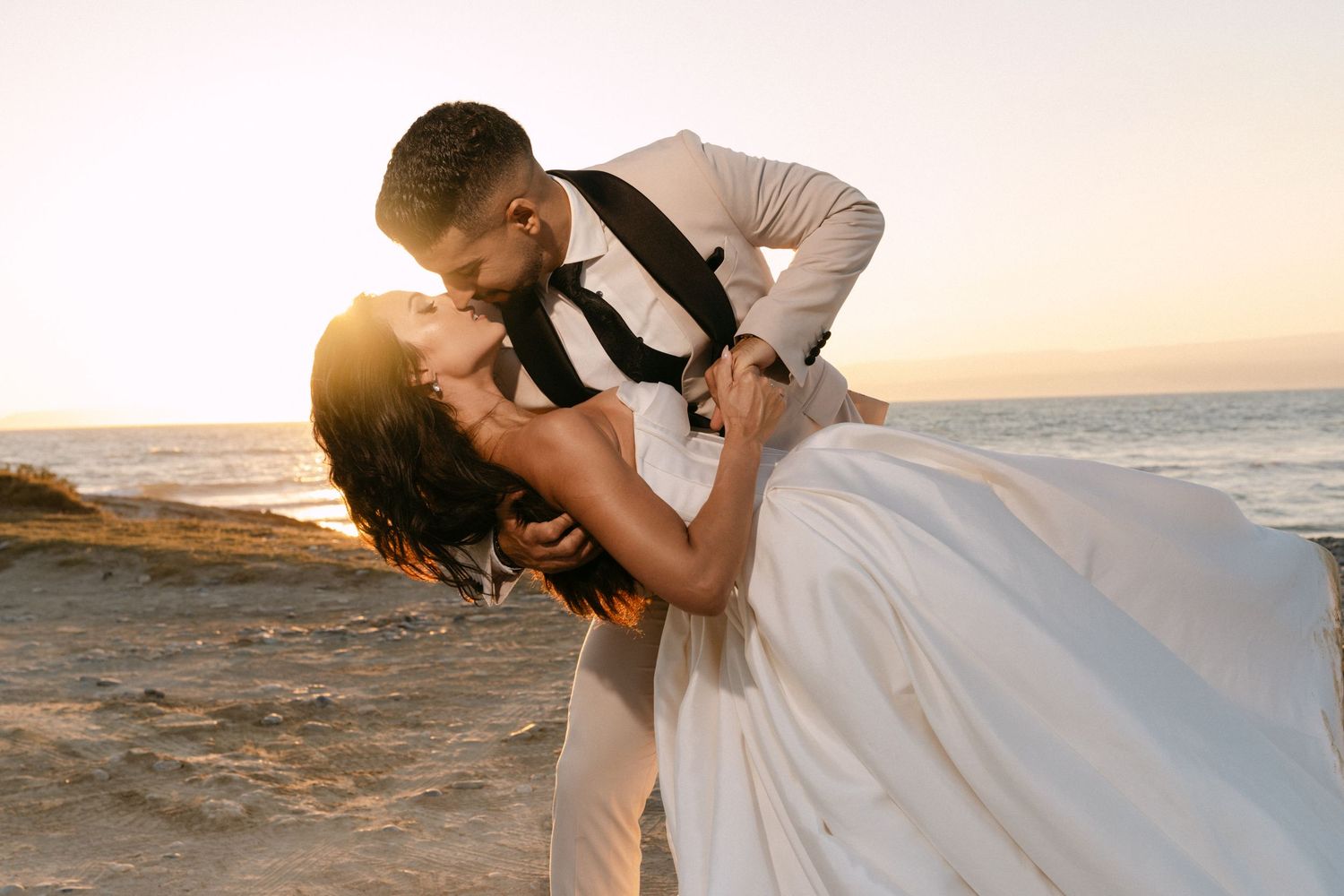 Groom dips bride in romantic beach kiss at golden sunset by the ocean.