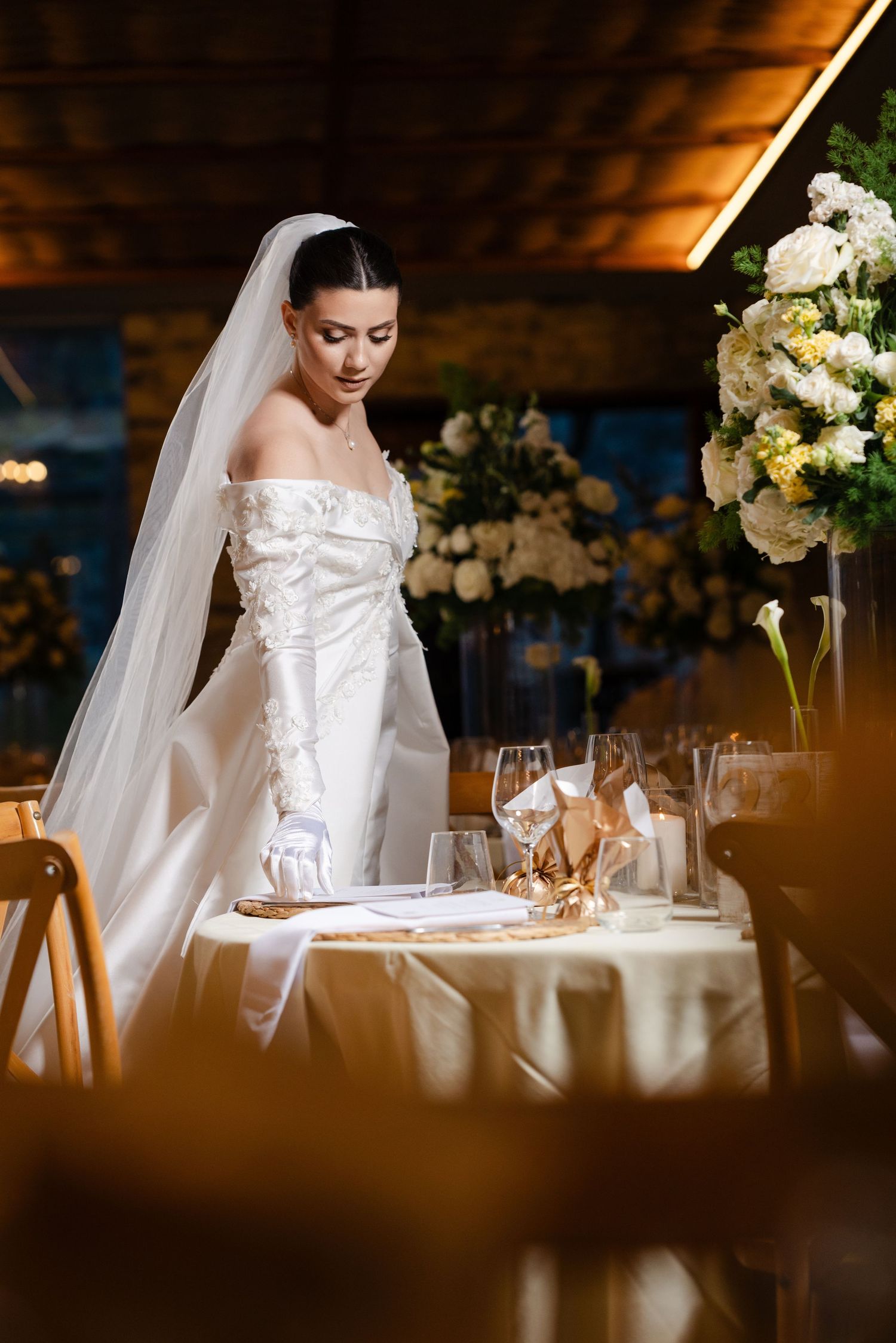 Elegant bride in off-shoulder white gown and gloves reviews table settings at decorated wedding reception.