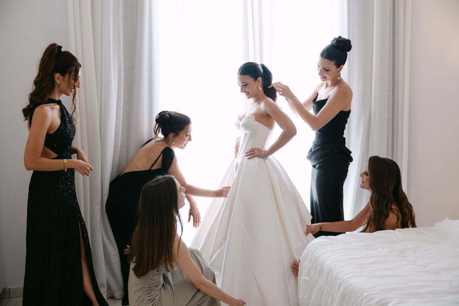 Bridesmaids in black dresses helping bride into white ball gown near bright window.
