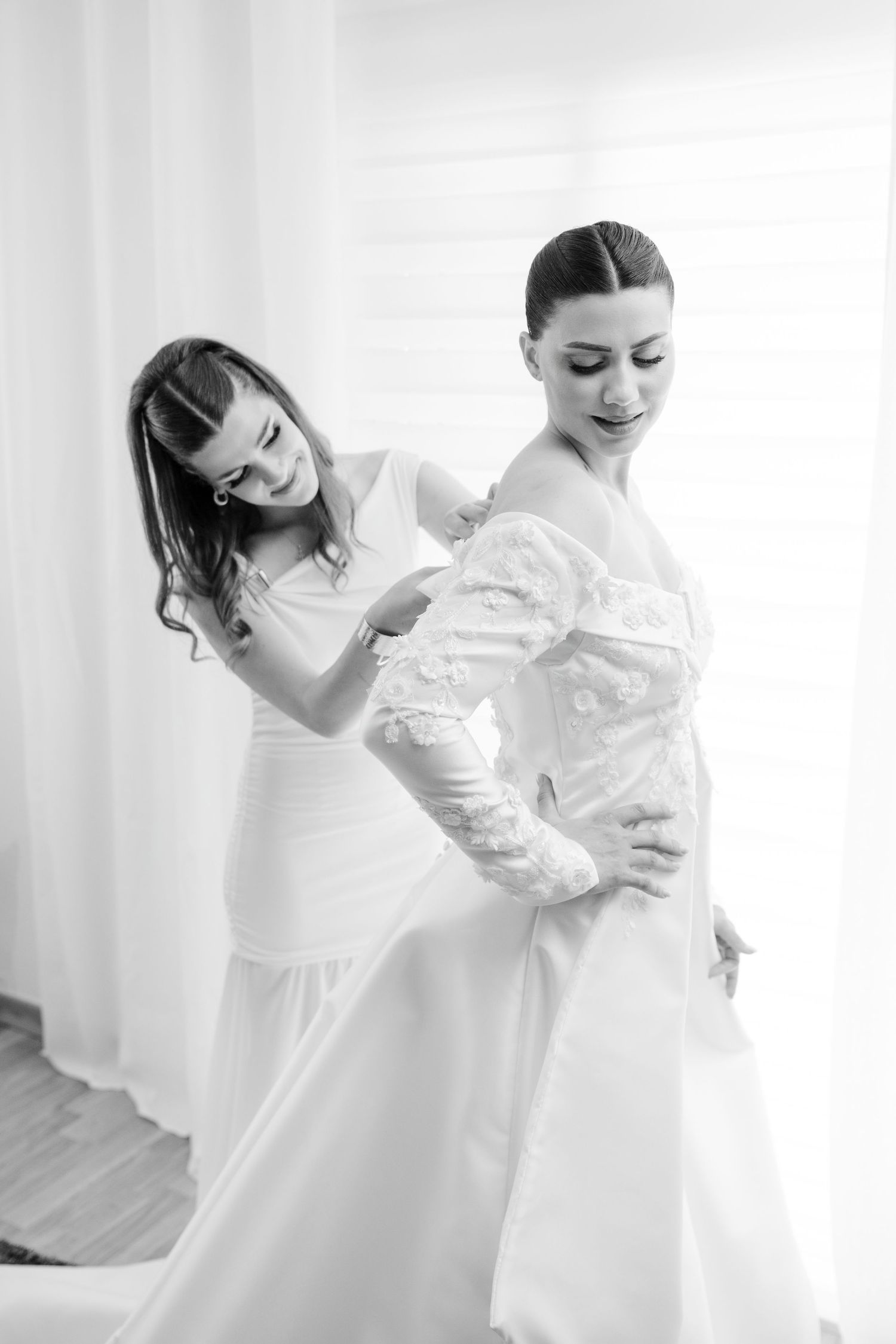 Black and white photo of bridesmaid helping bride button up elegant lace wedding gown.