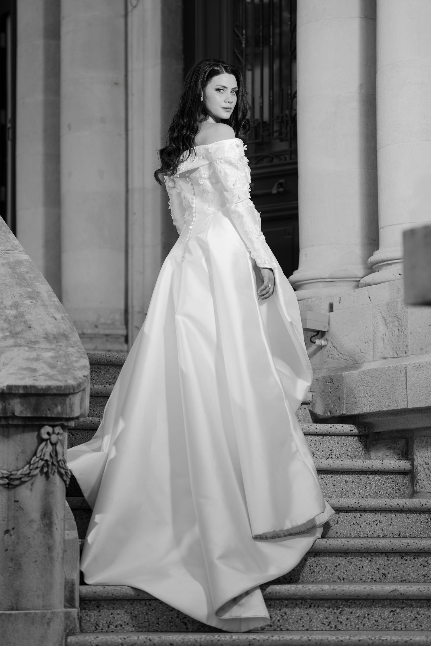 Black and white photo of a bride in a flowing white gown posing on elegant stone stairs.