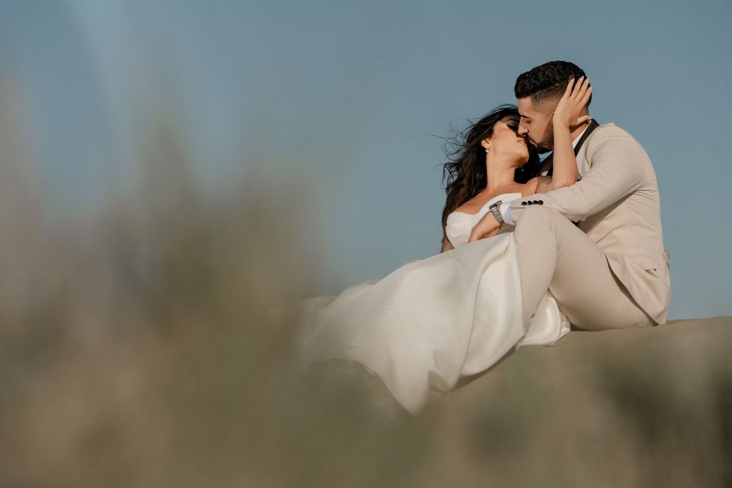 Bride and groom sit closely together about to kiss on a sandy dune under a soft blue sky.