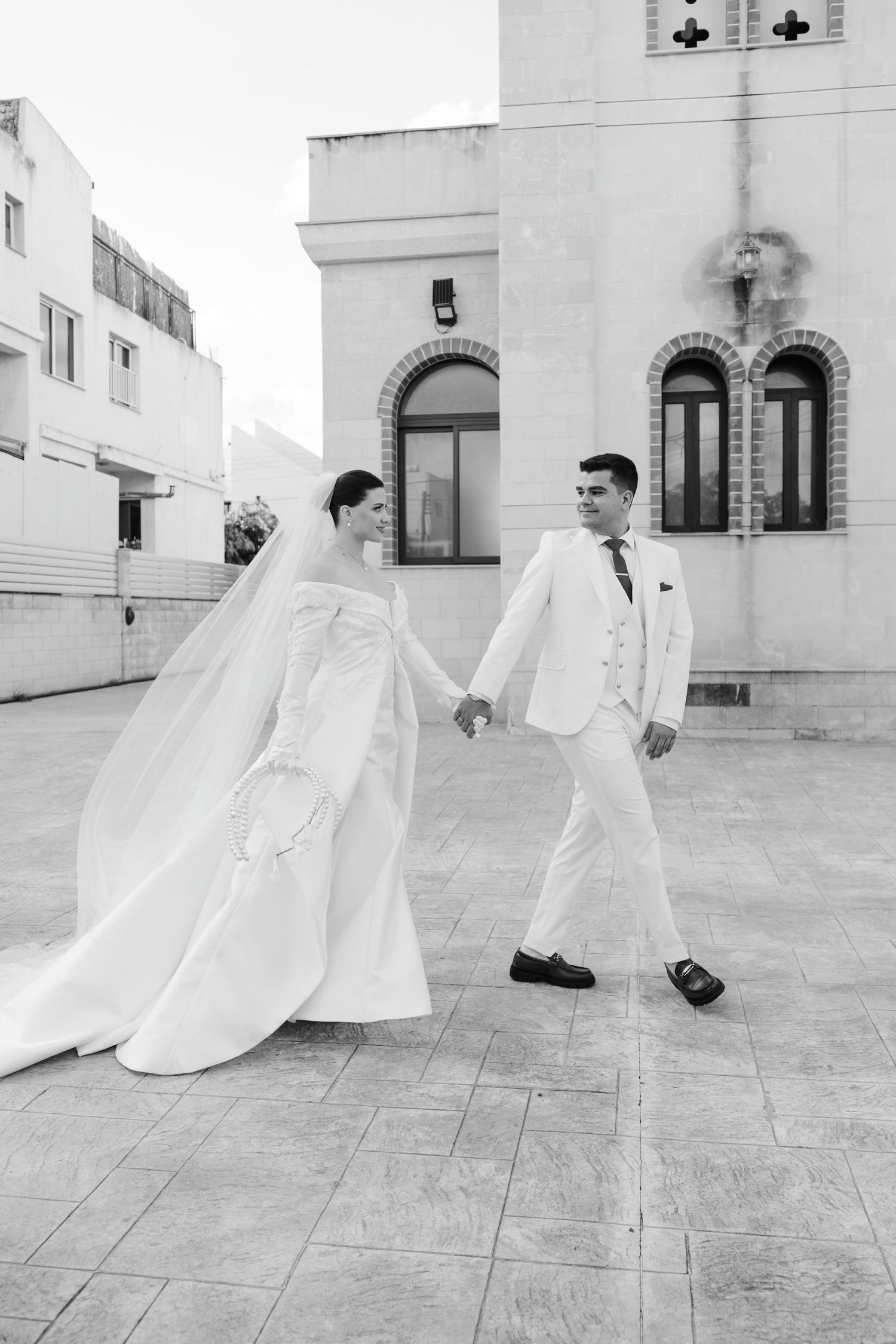 Bride and groom holding hands outside historic church in elegant white wedding attire.