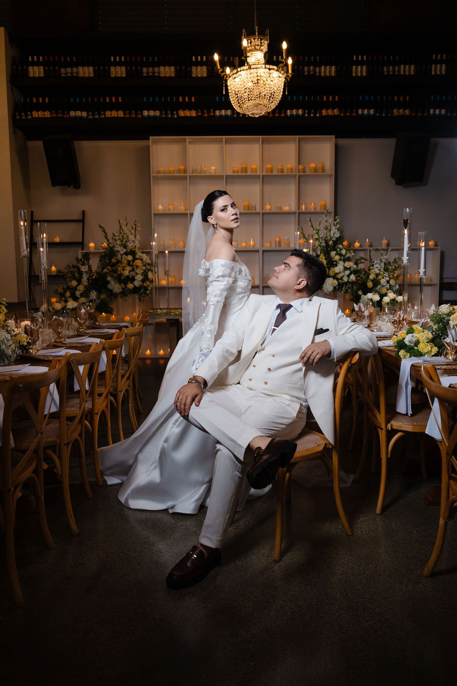 Bride and groom in coordinating white outfits pose beside candlelit reception tables under a crystal chandelier.
