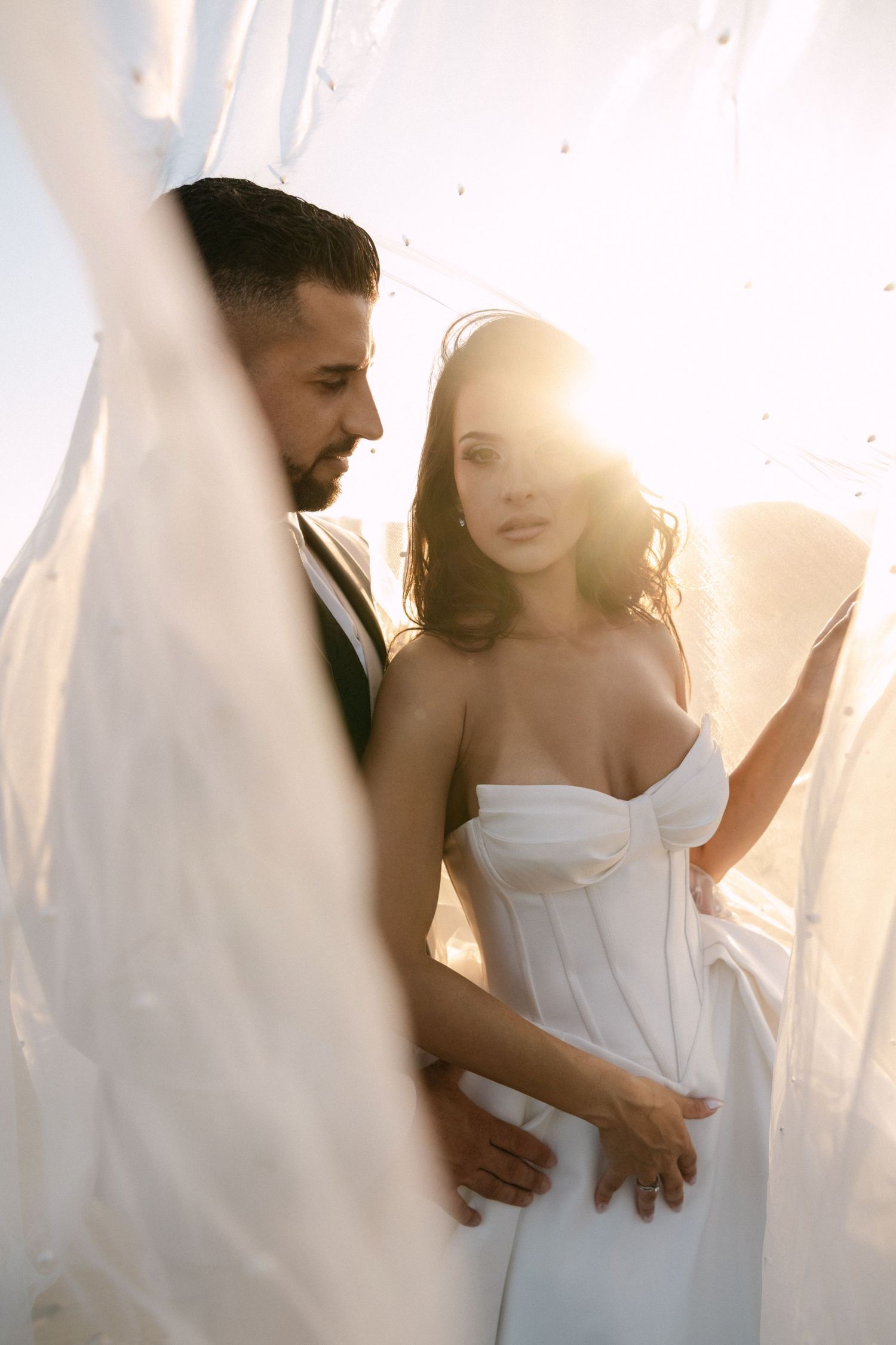 Bride and groom share an intimate moment surrounded by flowing white fabric in golden sunset light.