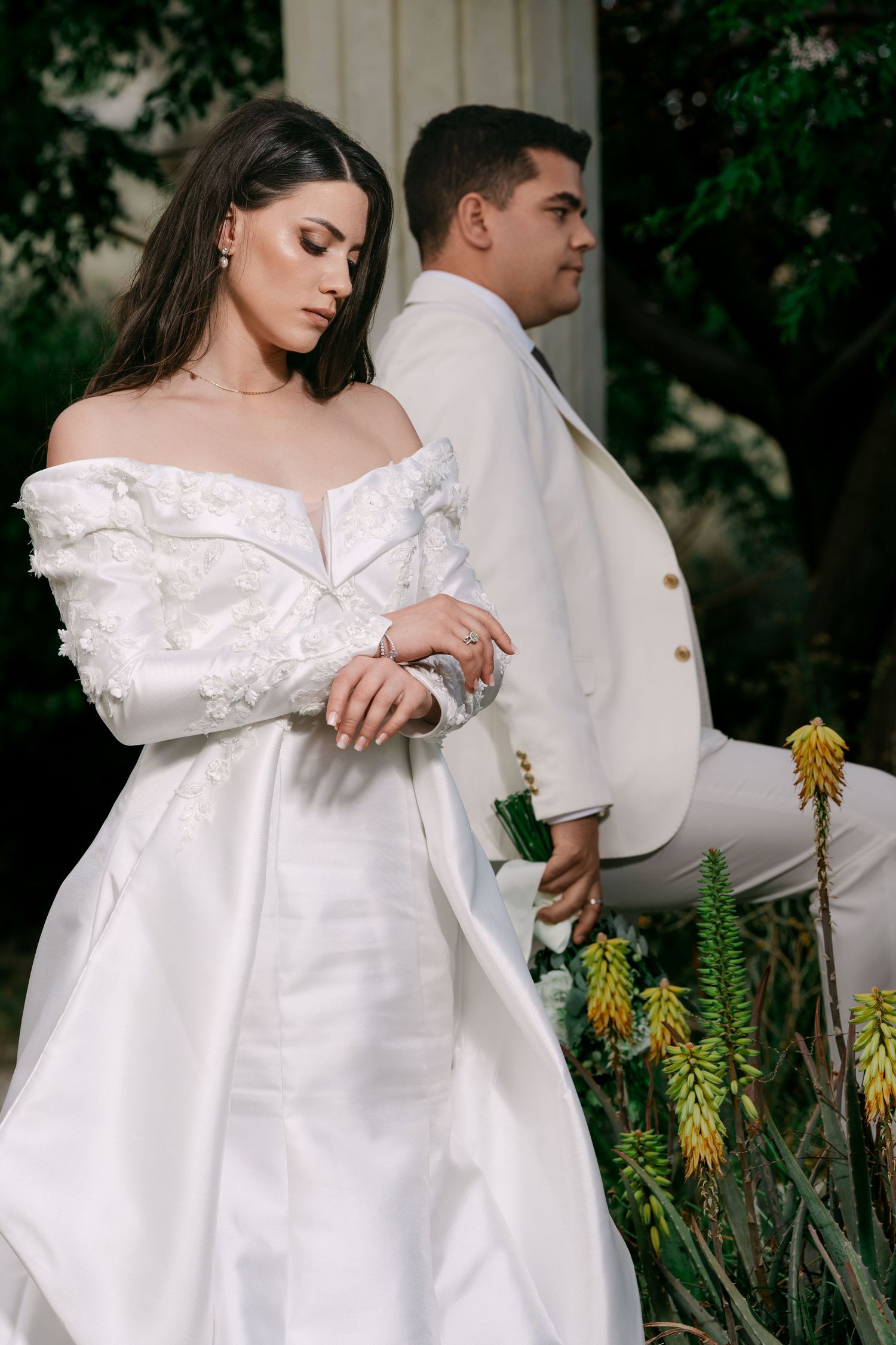 Elegant bride in white off-shoulder dress stands with groom in white suit amid tropical garden plants.