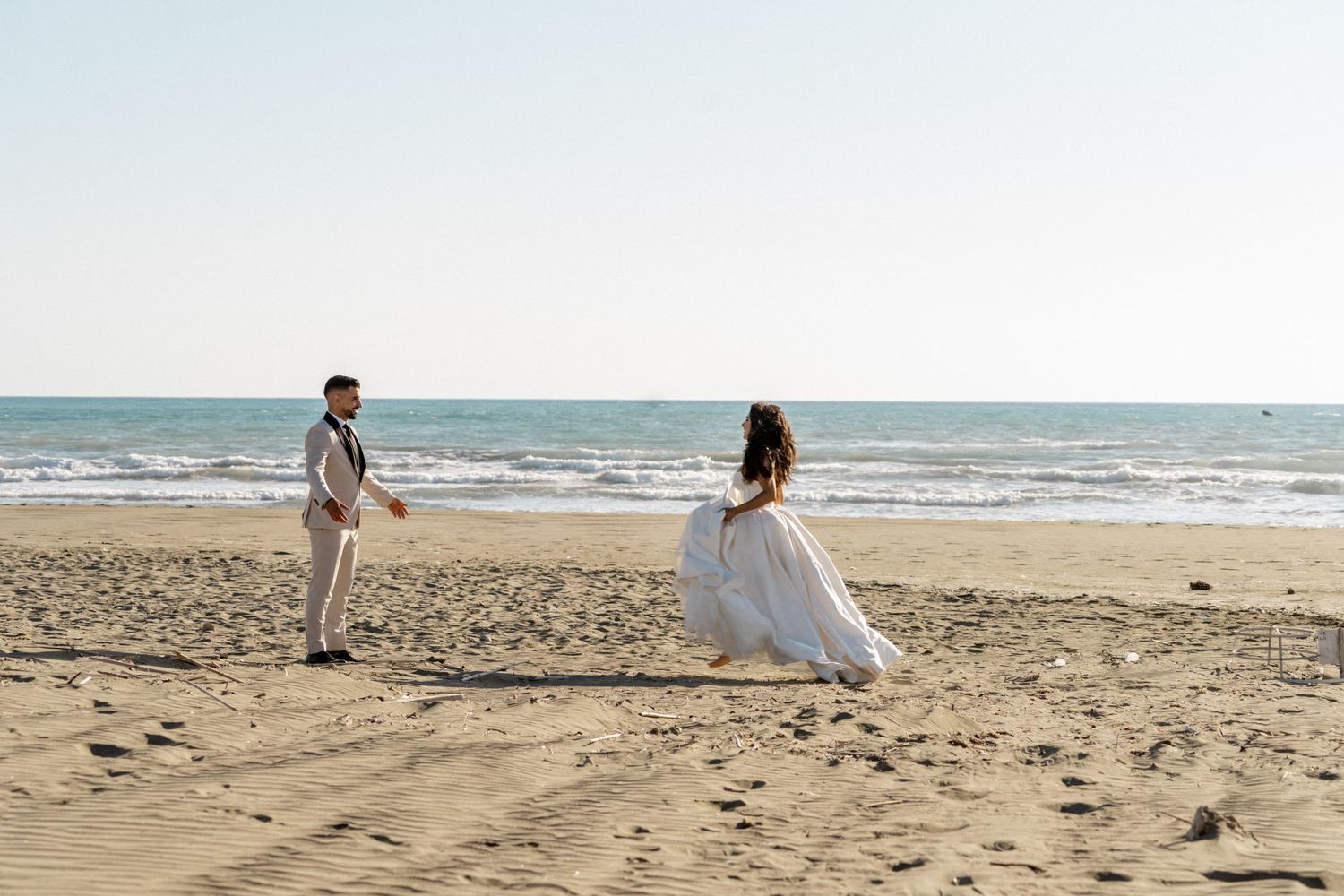 Bride in a white gown twirls toward groom on a sandy beach with ocean waves in the background.
