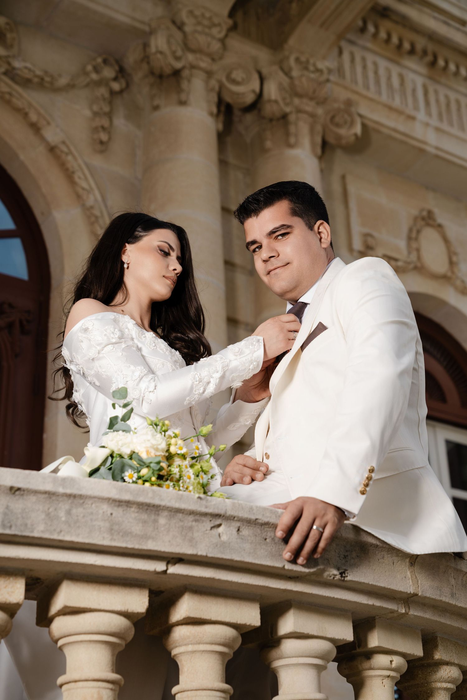 Bride adjusting grooms tie on a decorative balcony of a historic building on their wedding day.