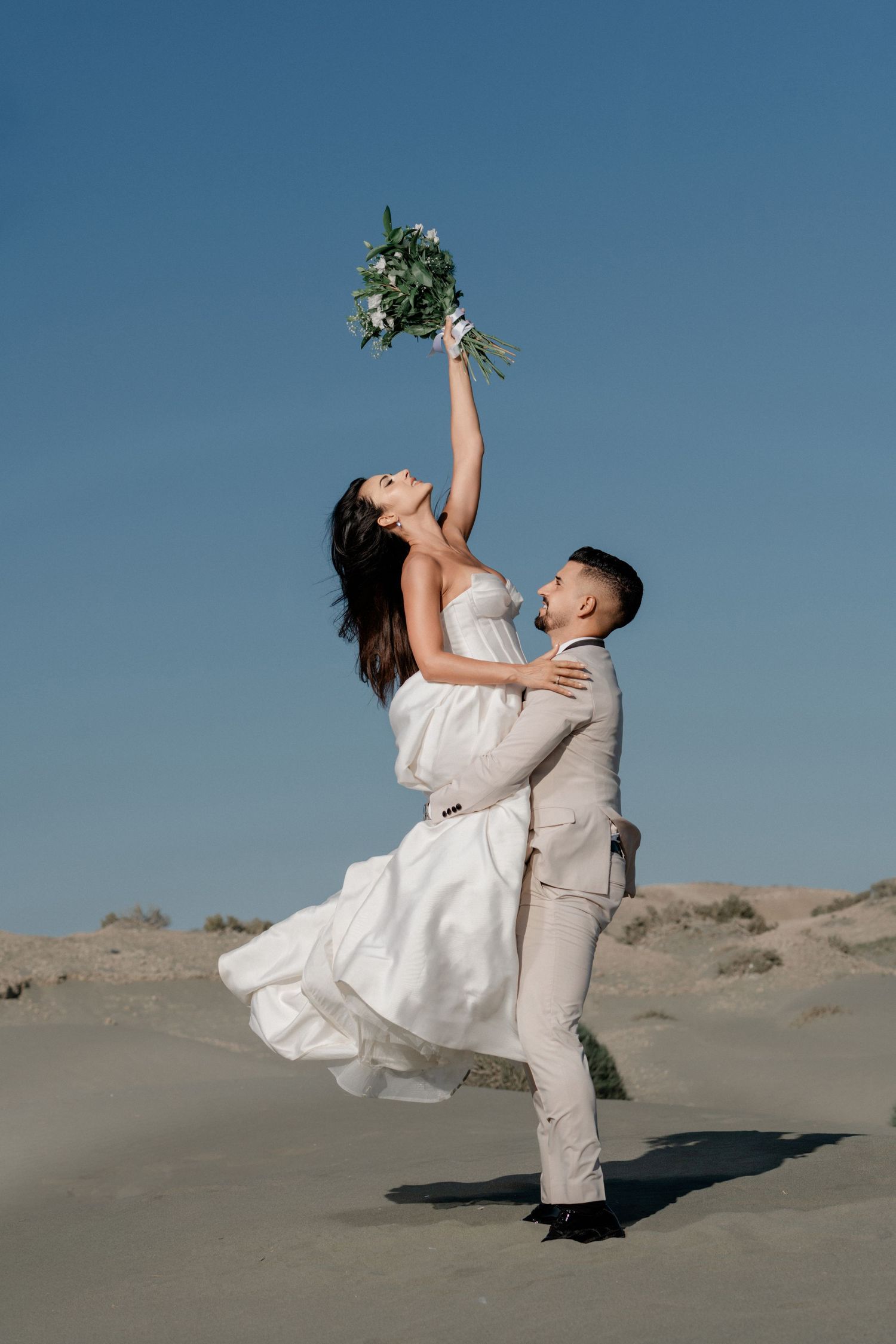 Groom dips bride as she raises a floral bouquet high on a sandy beach under a clear blue sky.