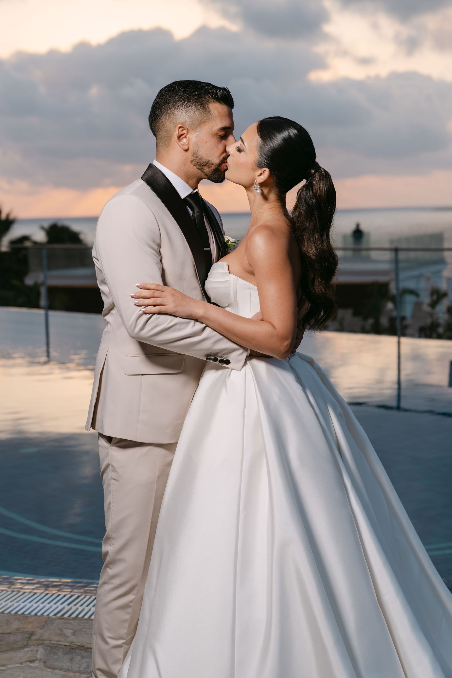 Bride and groom share a sunset kiss by an infinity pool in elegant wedding attire.