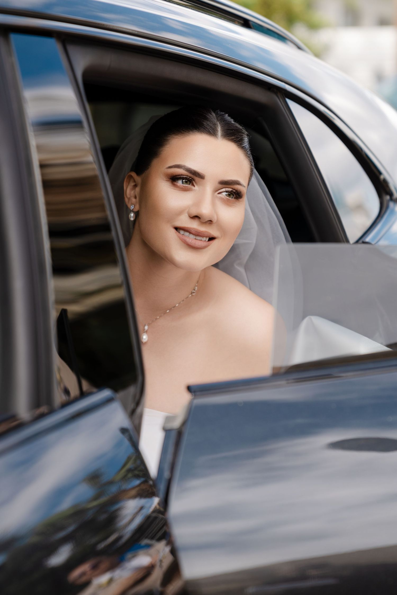 Beautiful bride in white veil and gown stepping out of luxury black car, smiling.