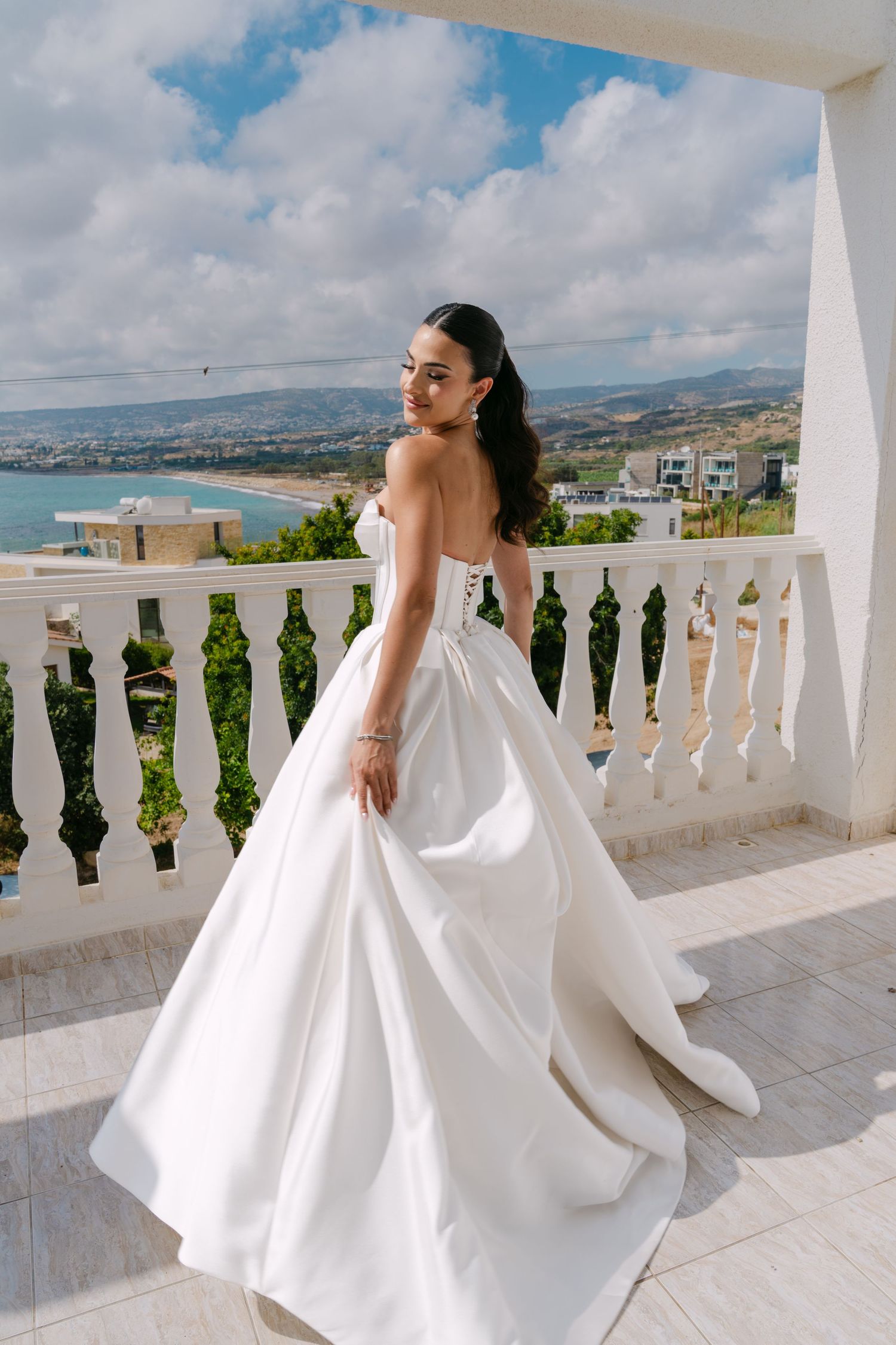 Bride in white strapless ball gown twirling on balcony with ocean backdrop.