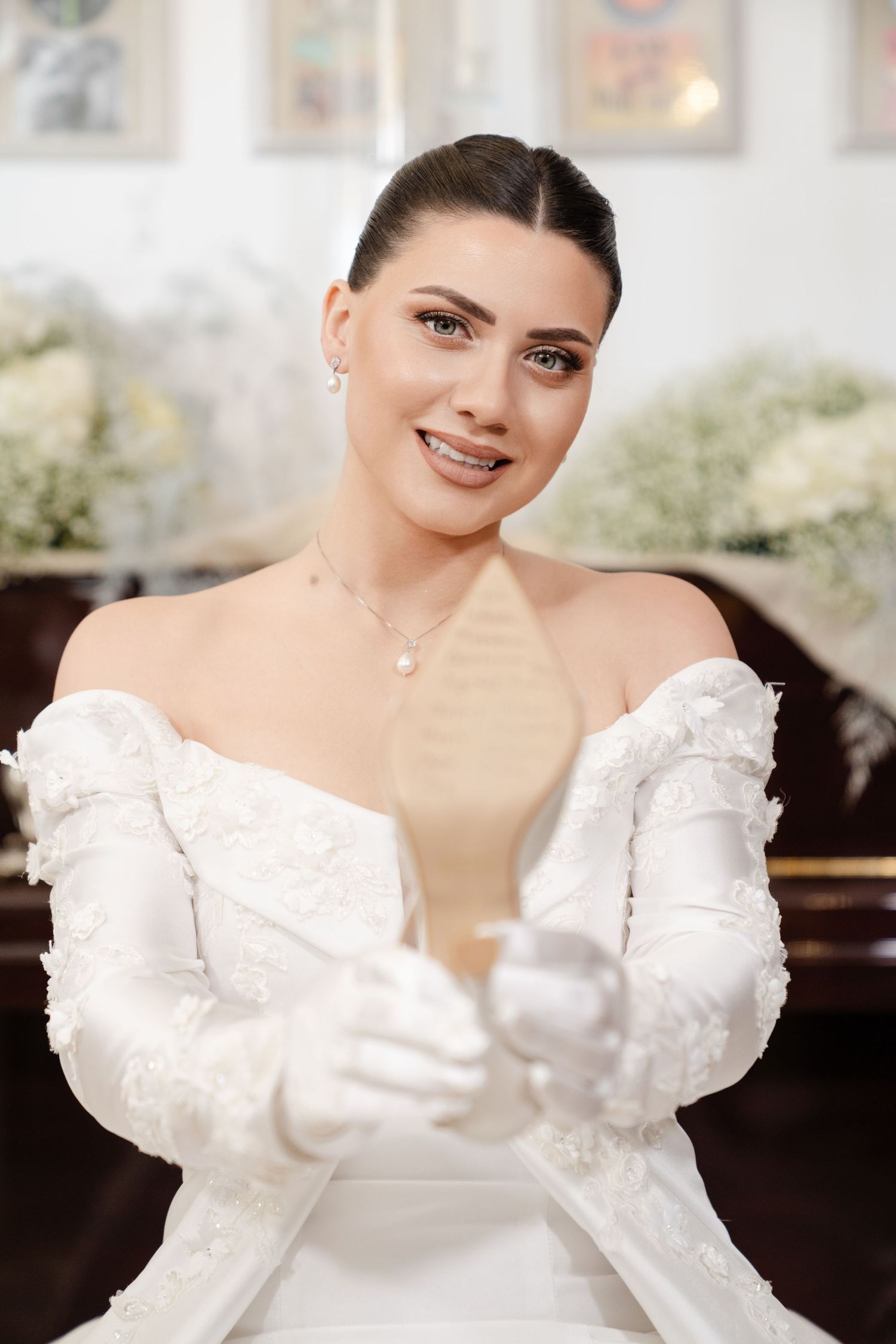 Smiling bride in off-shoulder lace gown and white gloves holding candle in church.