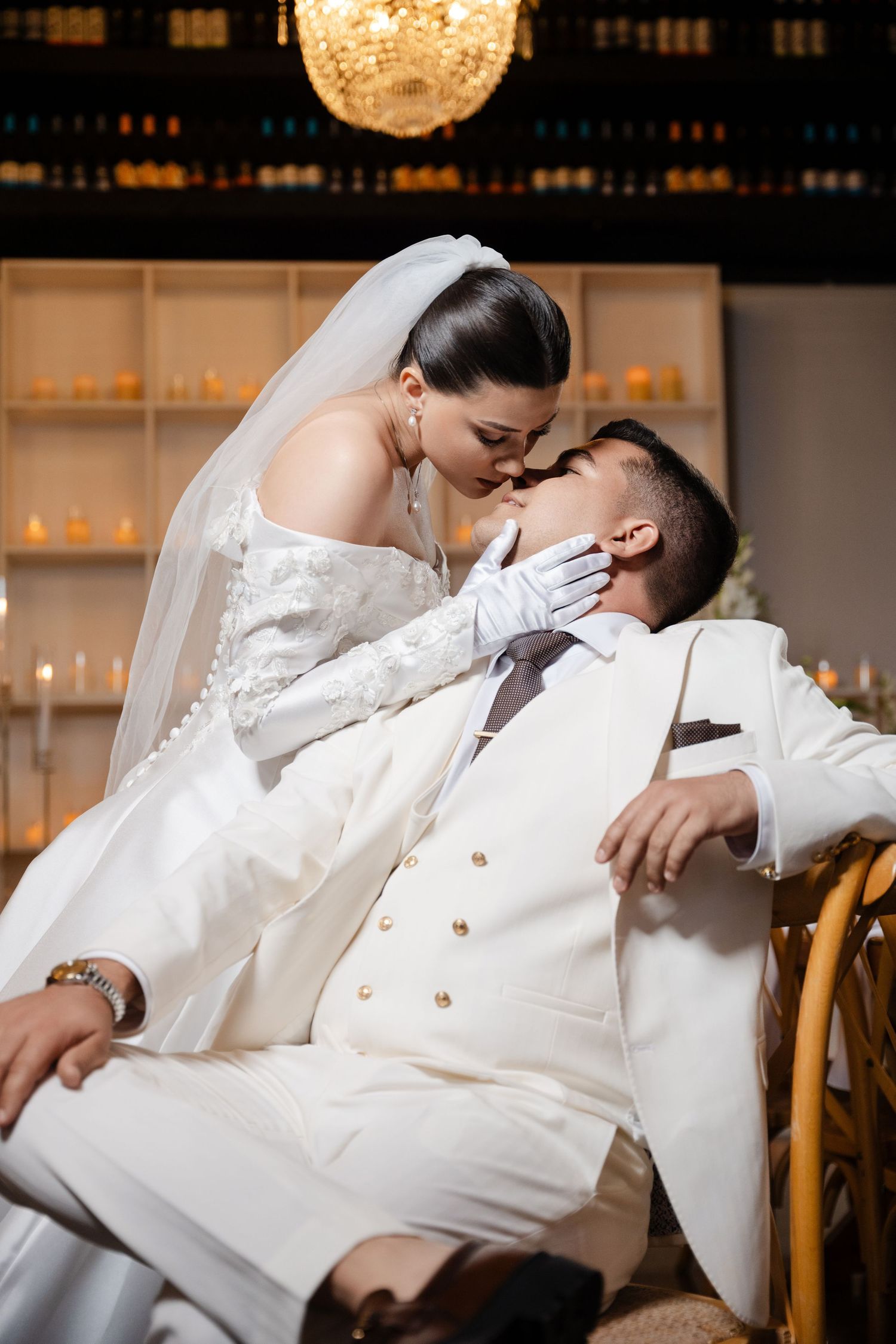 Bride in white gown and veil kisses groom in white suit while seated in elegant candlelit wedding venue.