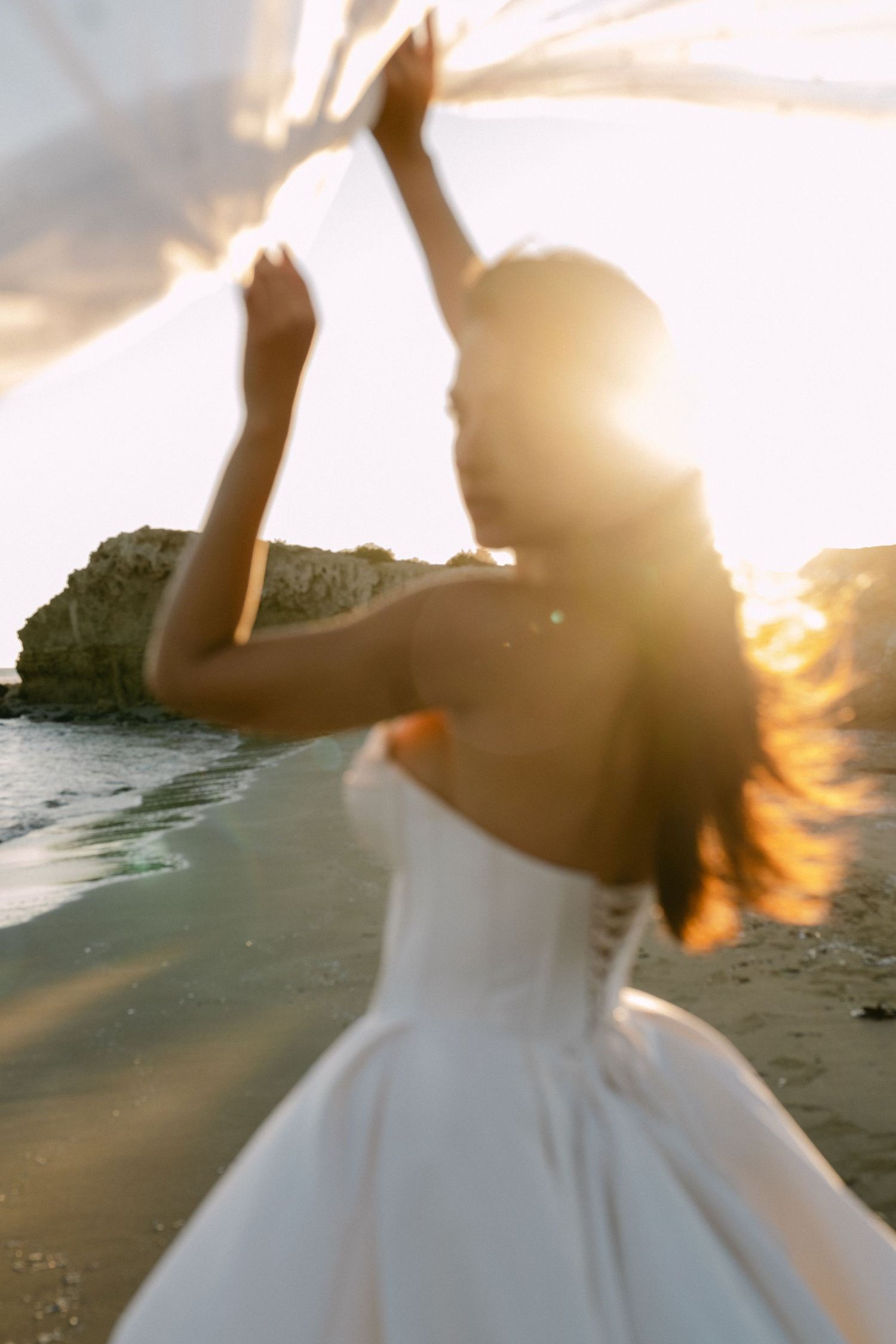 Bride in white strapless gown holds flowing veil on sandy beach during golden hour sunset.