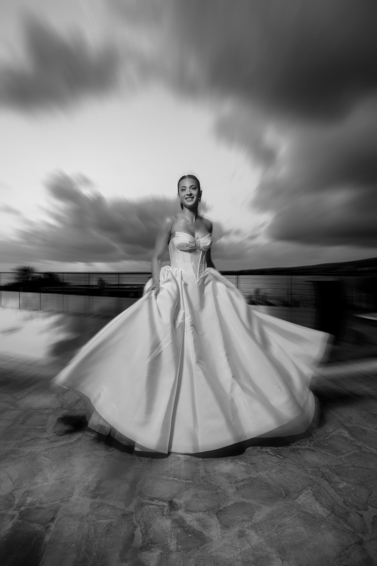 Bride twirling in strapless ball gown under dramatic cloudy sky in artistic motion blur photo.