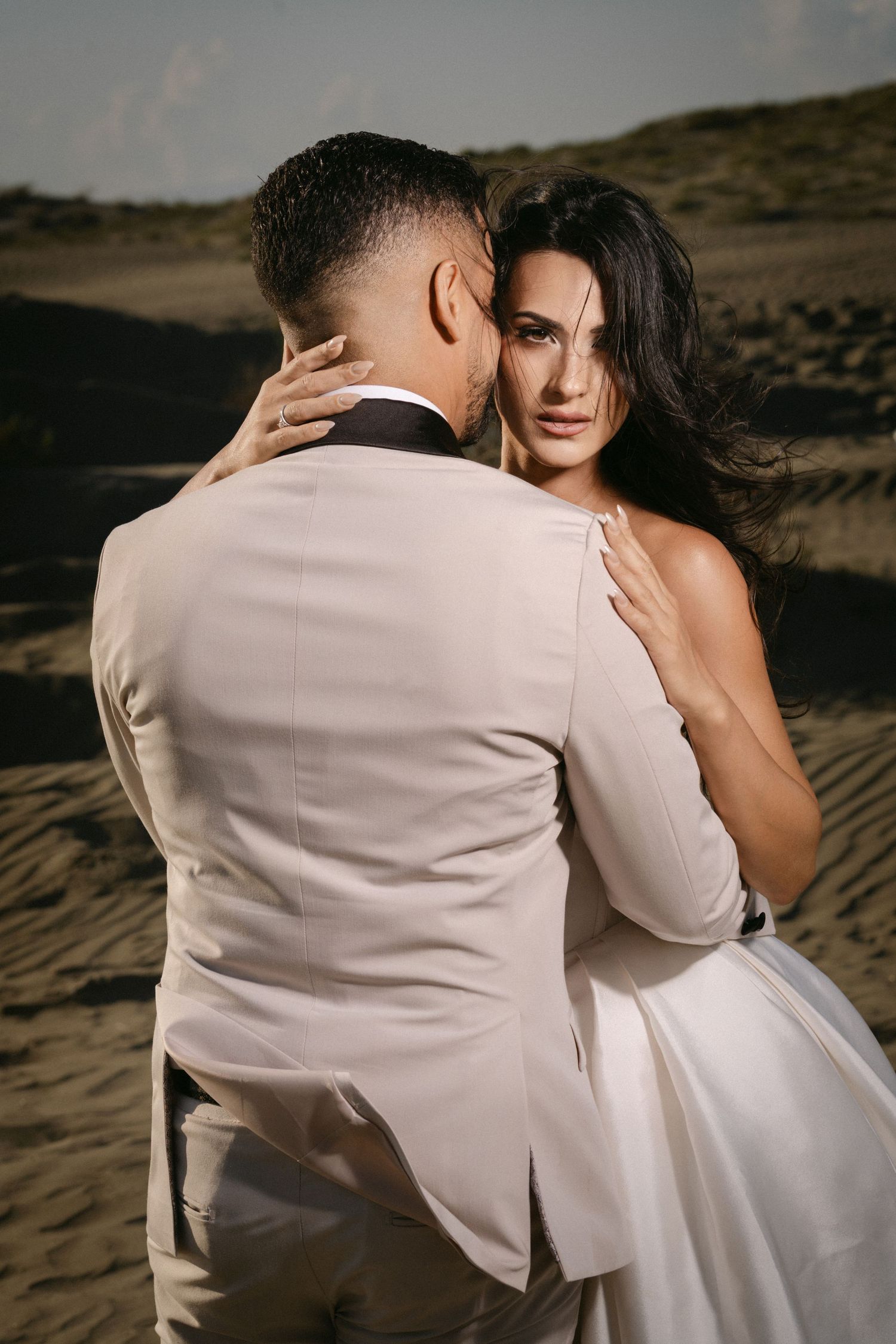 Bride and groom embrace romantically in sandy desert dunes, groom in beige tuxedo kissing brides cheek.