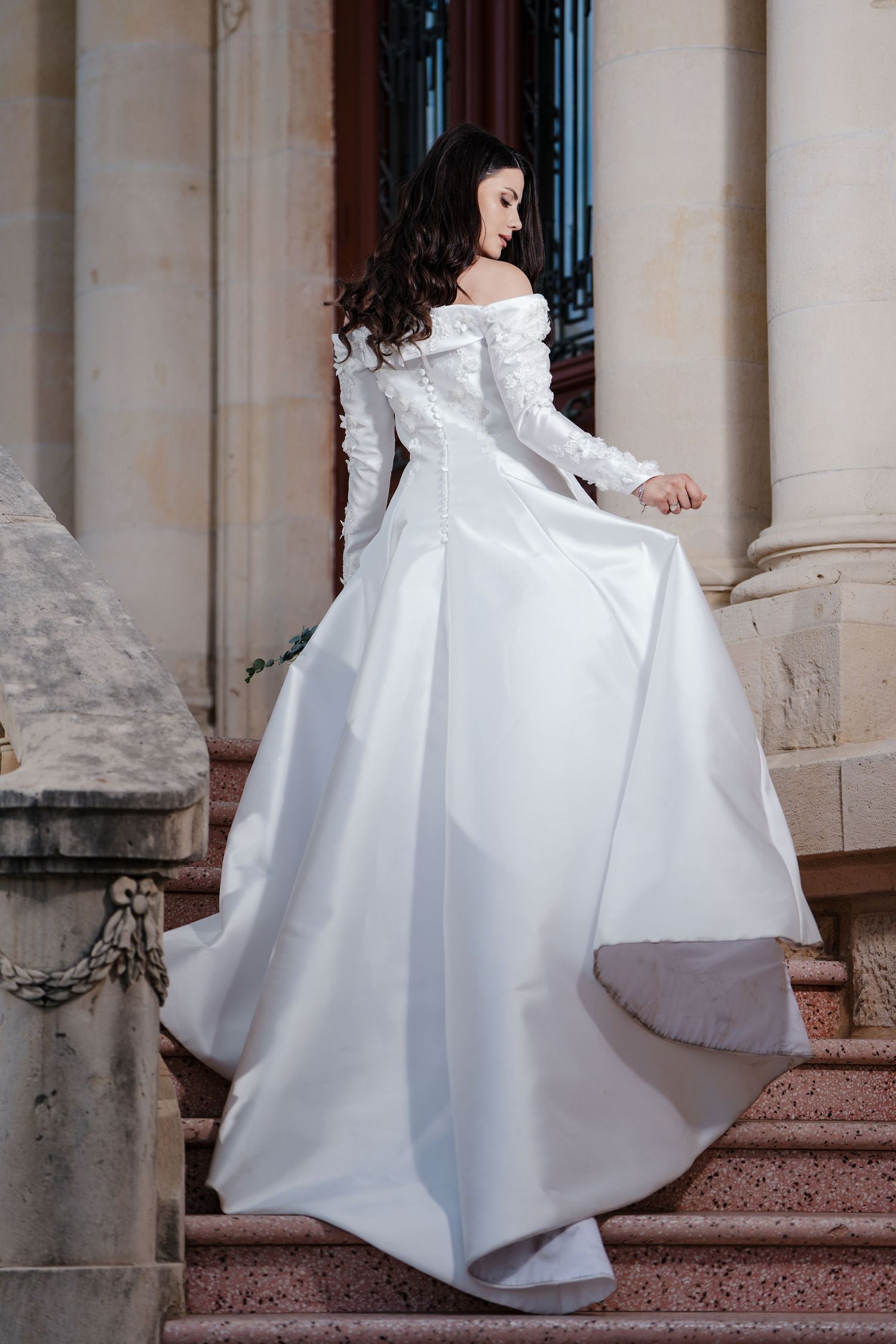 Bride in a long-sleeve white wedding dress elegantly lifting her skirt on ornate stone steps.