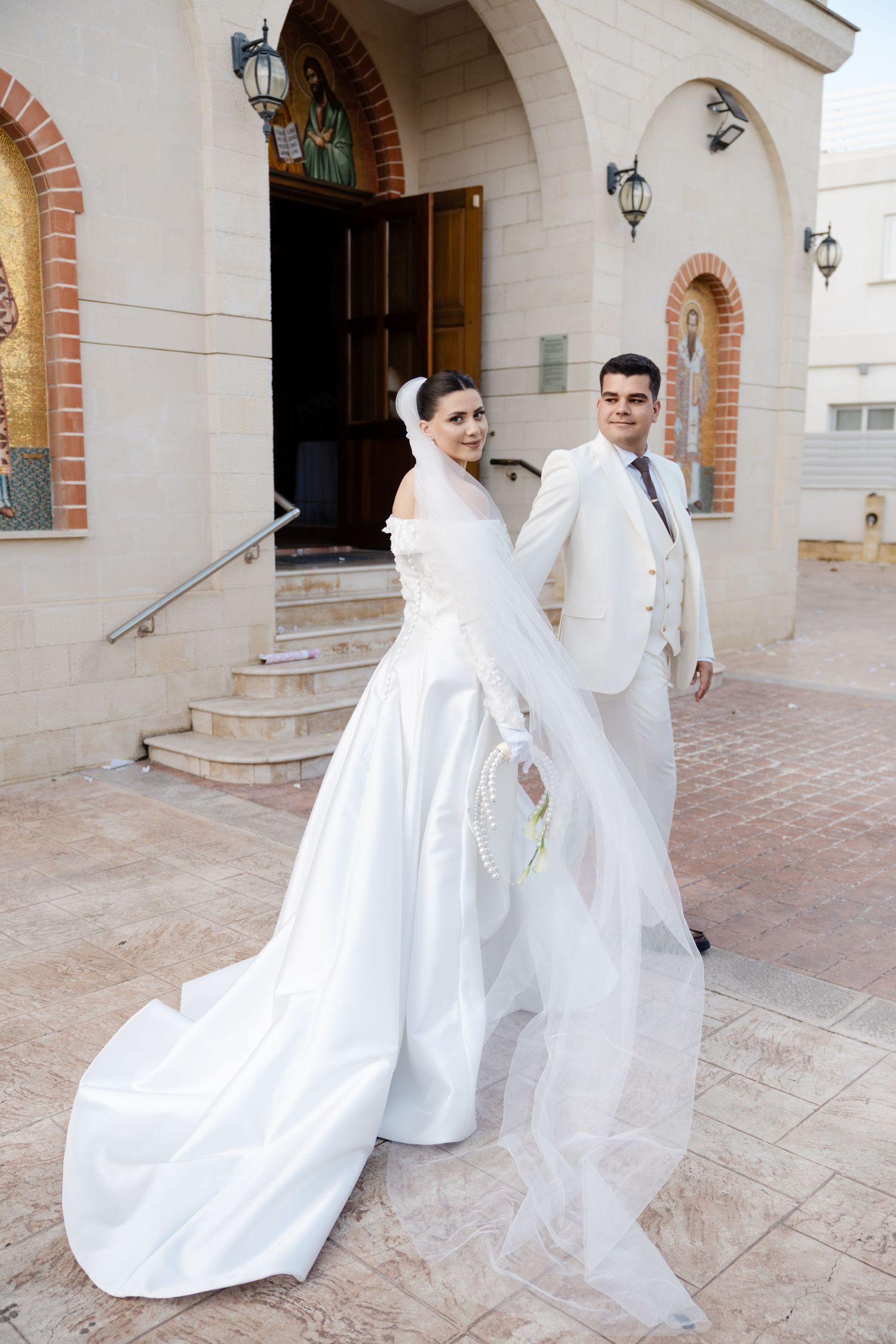 Bride in long white gown with veil and groom in white suit outside church.