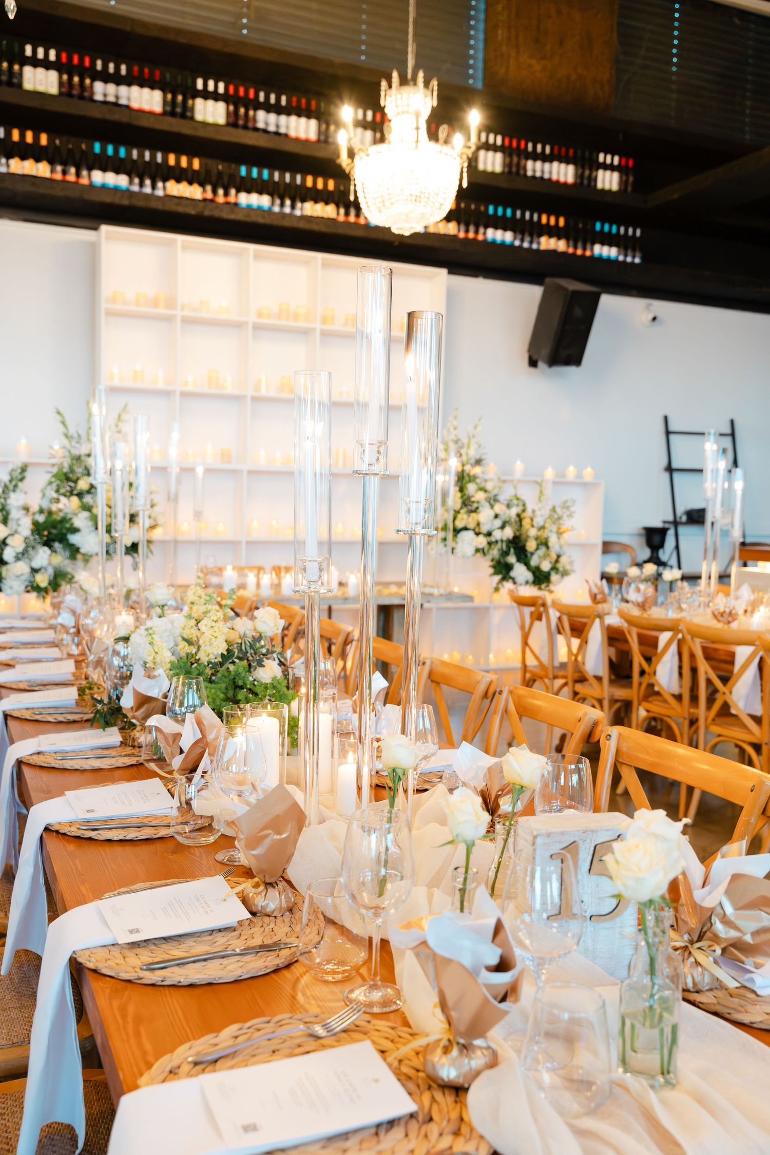 Luxurious wedding reception with long tables, candelabras, white florals, and a wine bottle display backdrop.