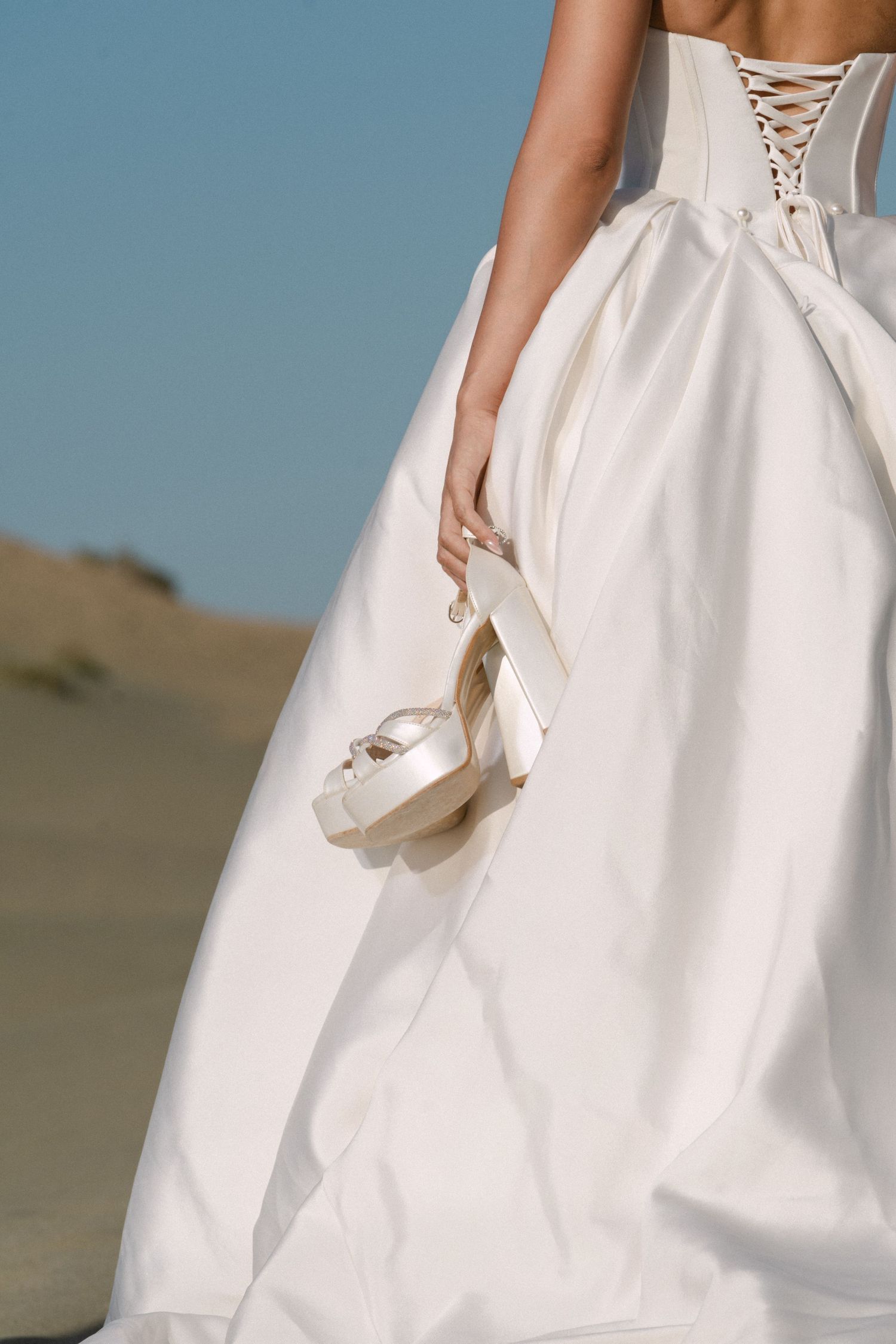 Close-up of brides white corset ballgown with lace-up back detail as she holds white heels in desert.