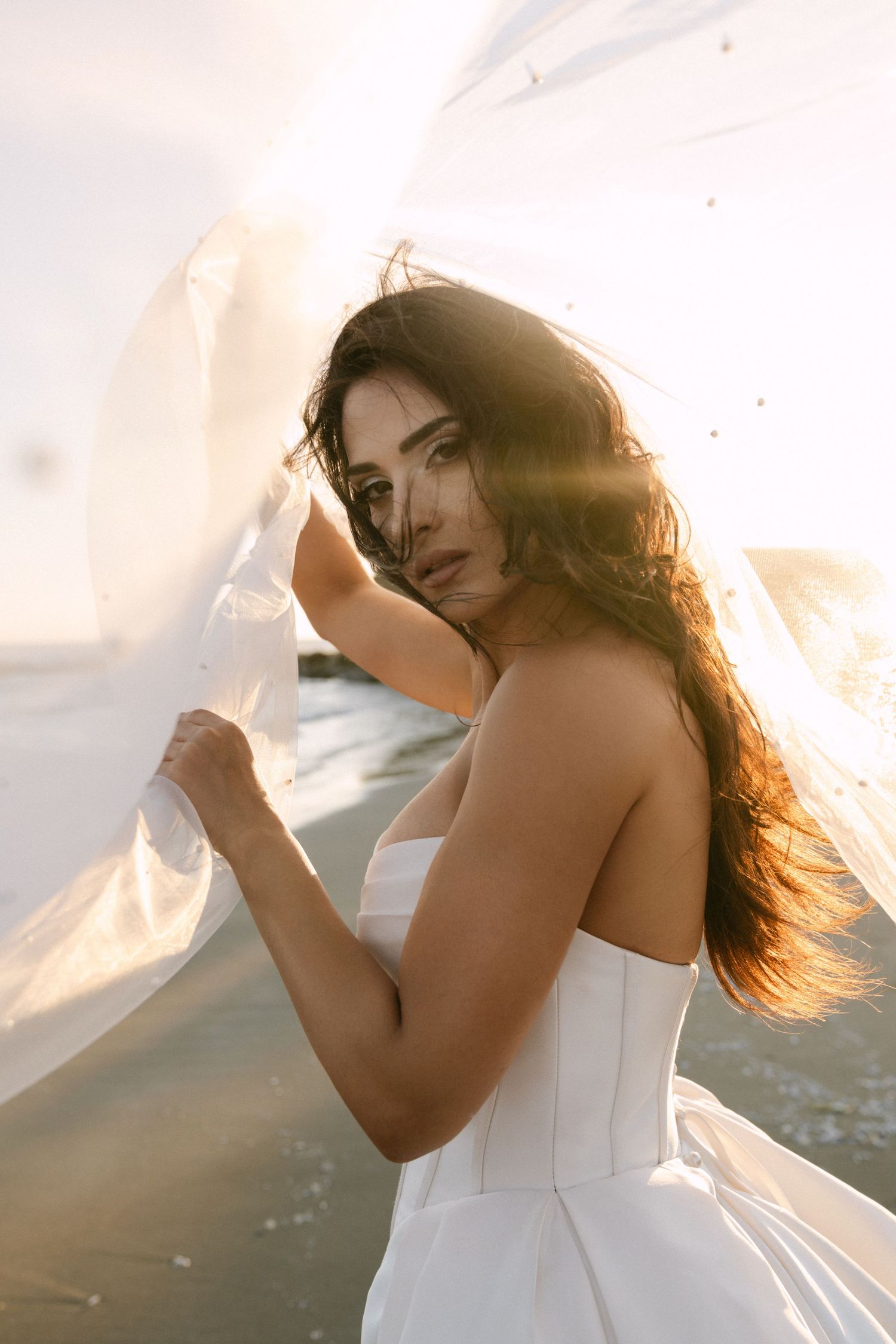 Beautiful dark-haired bride in white strapless gown holds flowing veil at beach sunset.