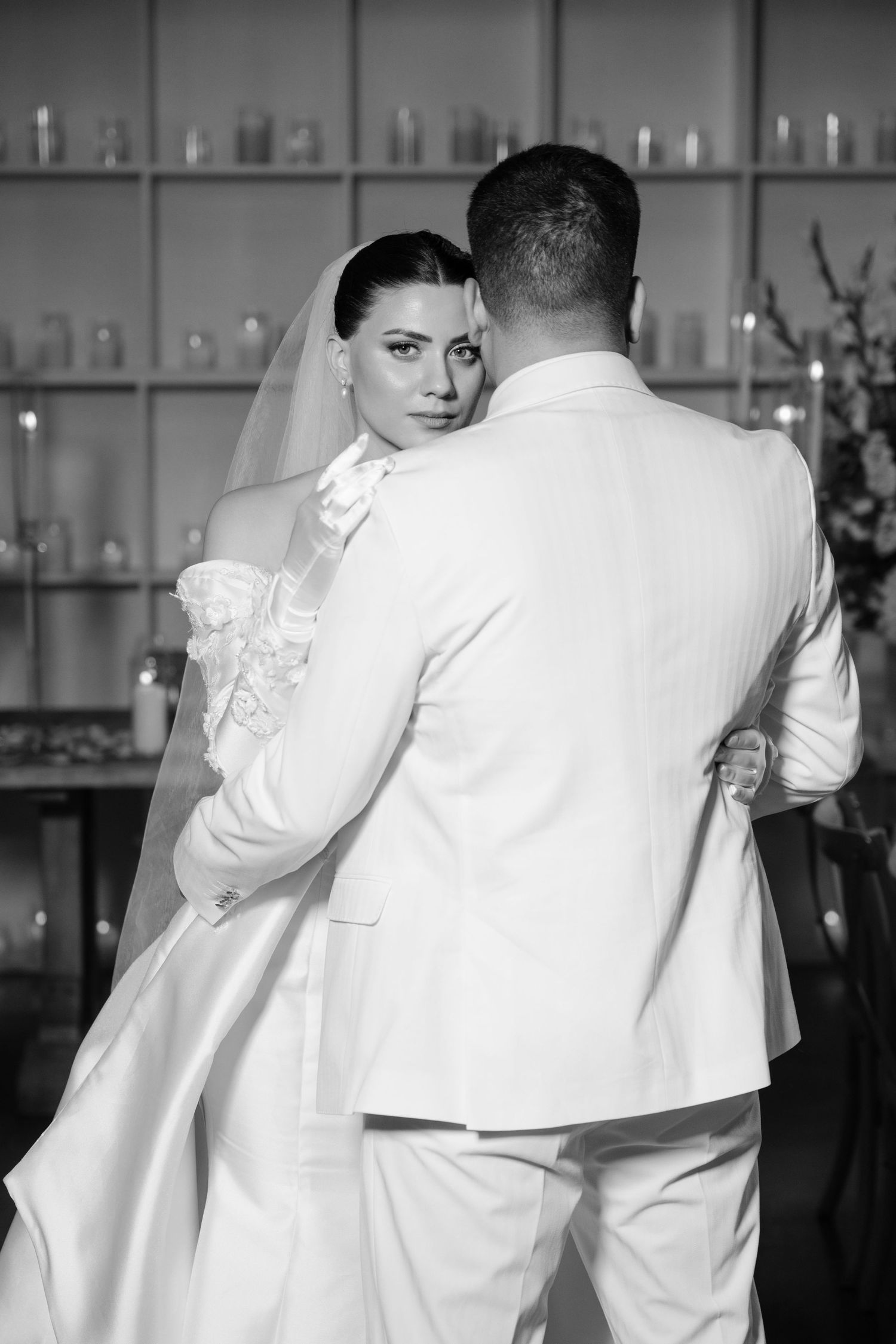 Black and white photo of bride in veil embracing groom from behind in white suit at wedding reception.