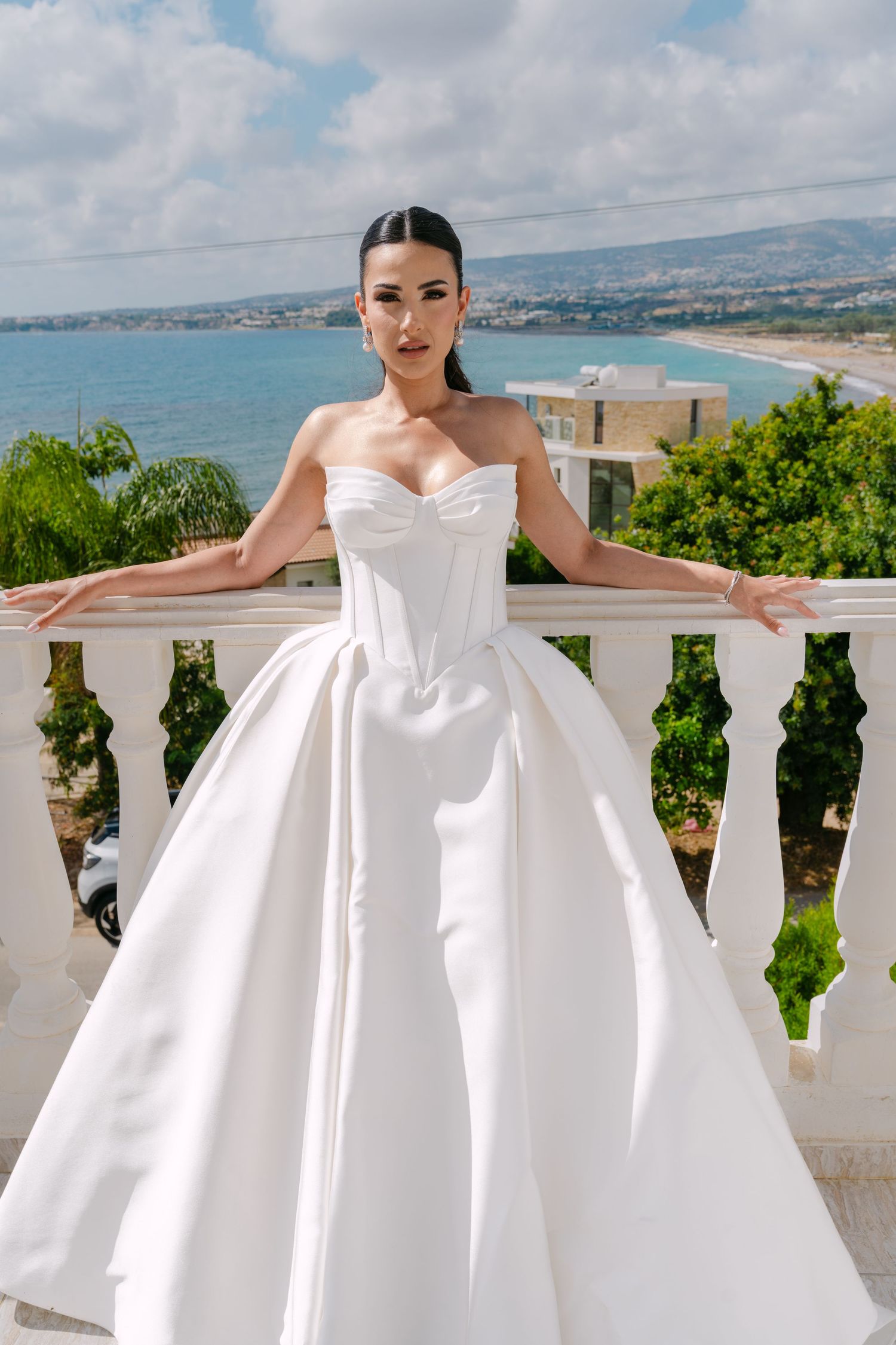 Elegant bride in strapless white ball gown posing on balcony with coastal view.