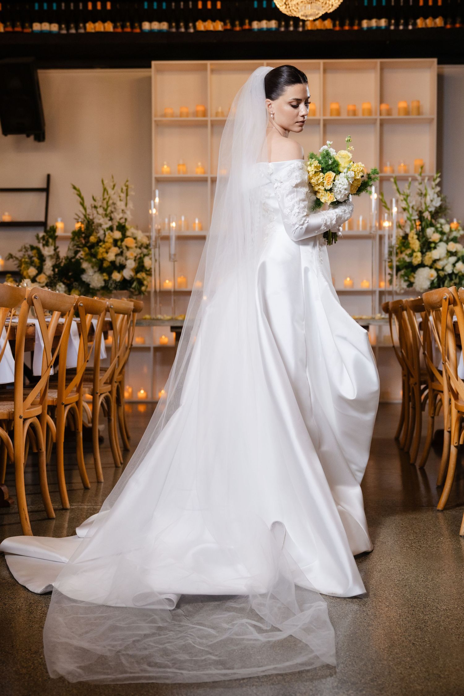 Bride in white off-shoulder gown holds yellow and white bouquet beside candlelit reception tables with veil flowing.