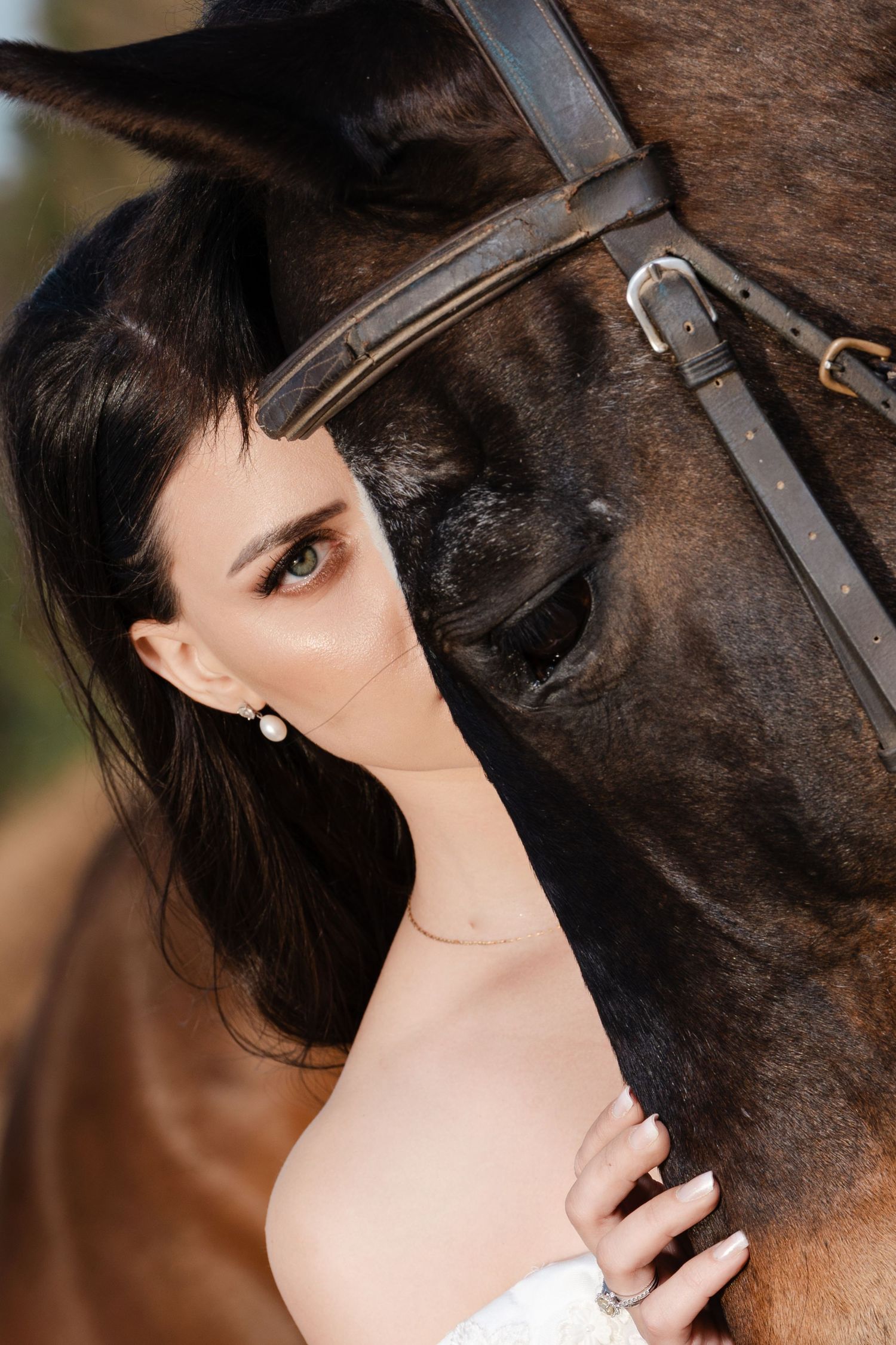 Close-up portrait of dark-haired bride in white dress posing beside a brown horse with bridle.