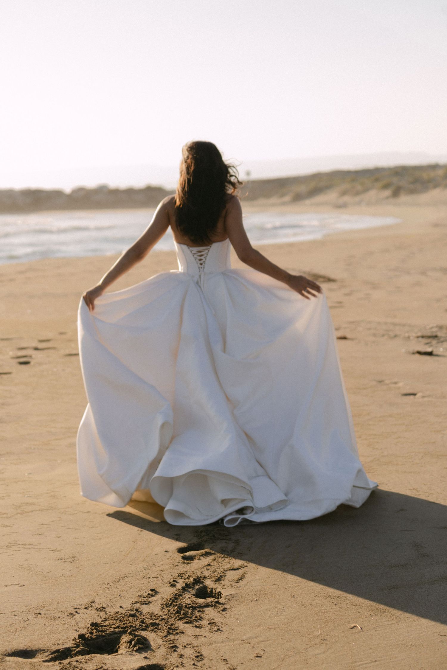 Bride in strapless white ballgown with lace-up back walks along sandy beach with arms outstretched.