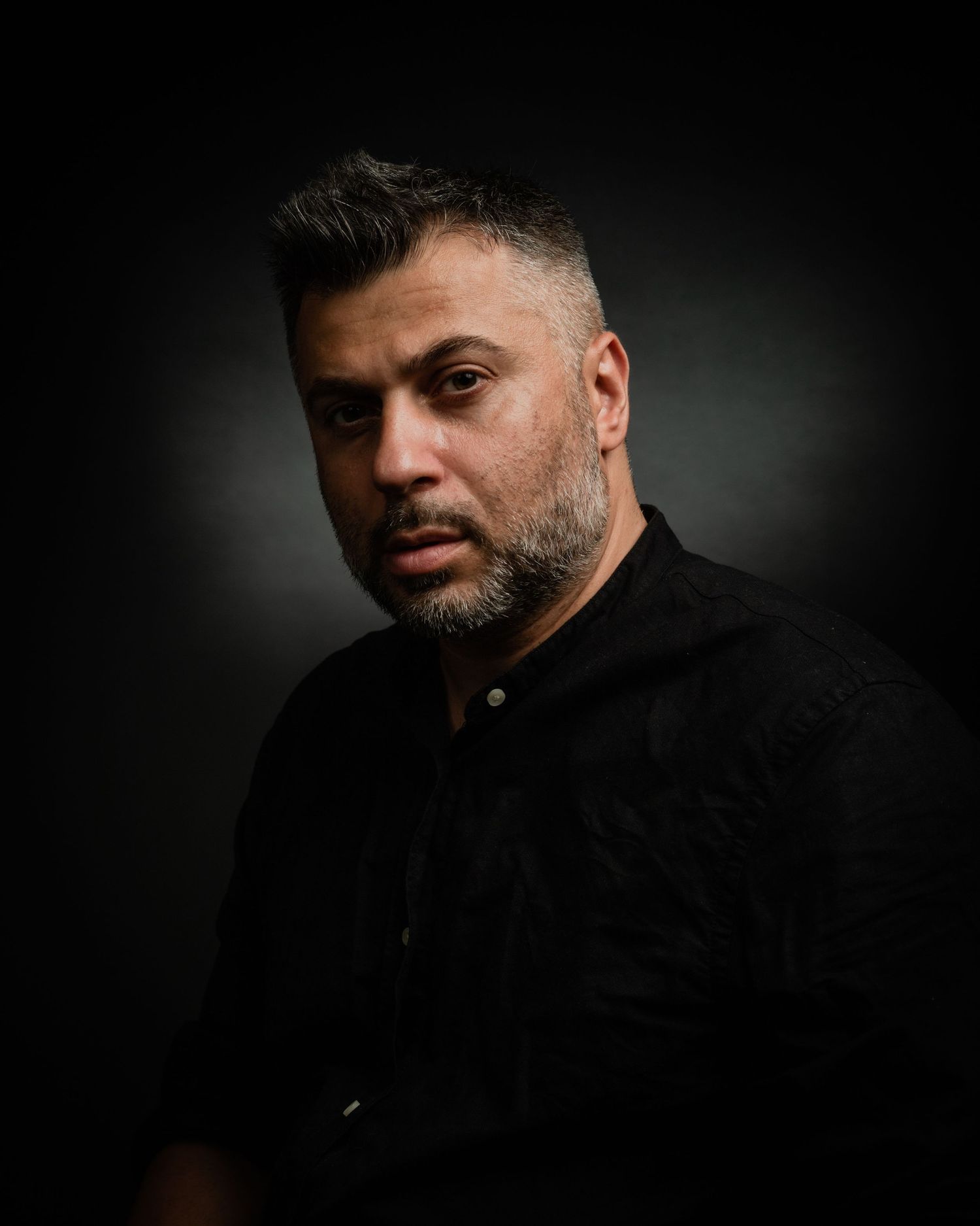 Man with short hair and beard wearing a black shirt in a dramatic dark studio portrait.