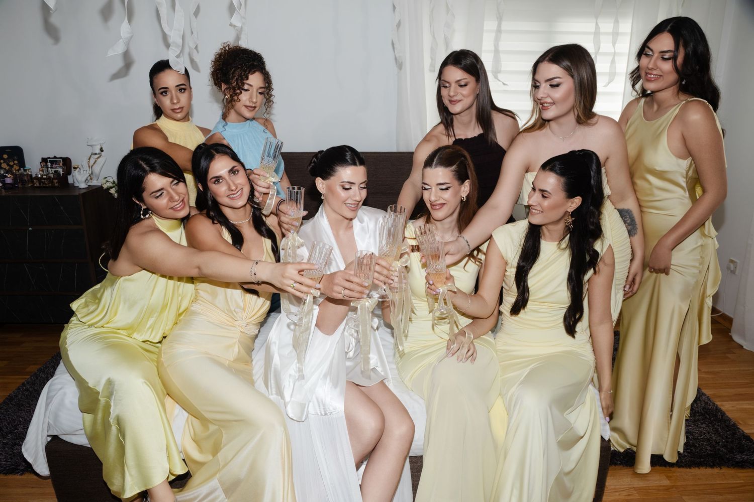Bride in white robe celebrating with bridesmaids in yellow dresses, toasting with champagne.