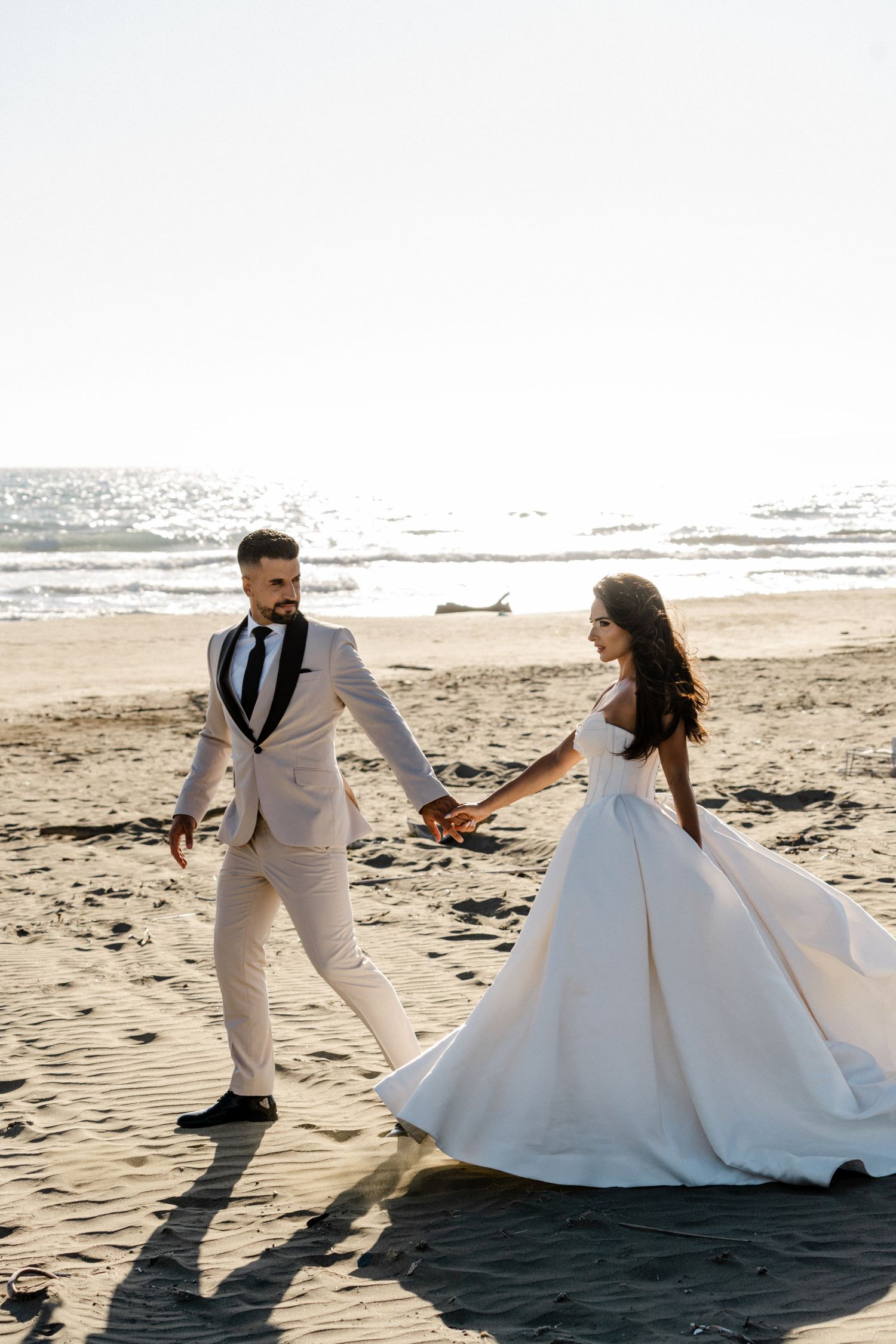 Bride and groom hold hands walking along a sunlit beach, she in a white gown, he in a cream suit.