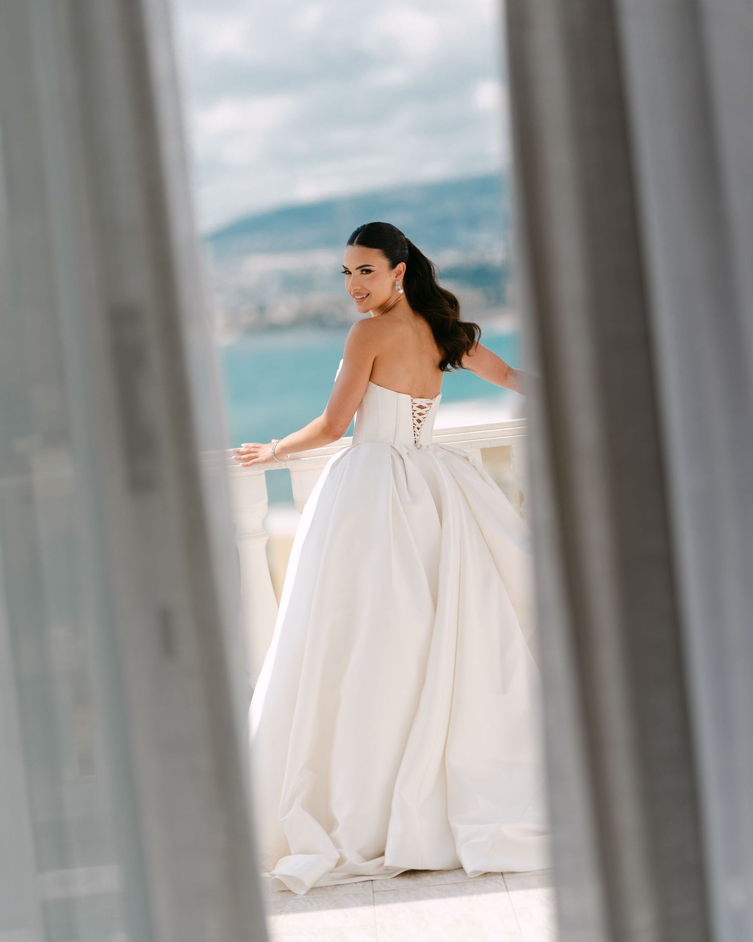 Bride in strapless ball gown with corset back stands on balcony overlooking scenic ocean view.