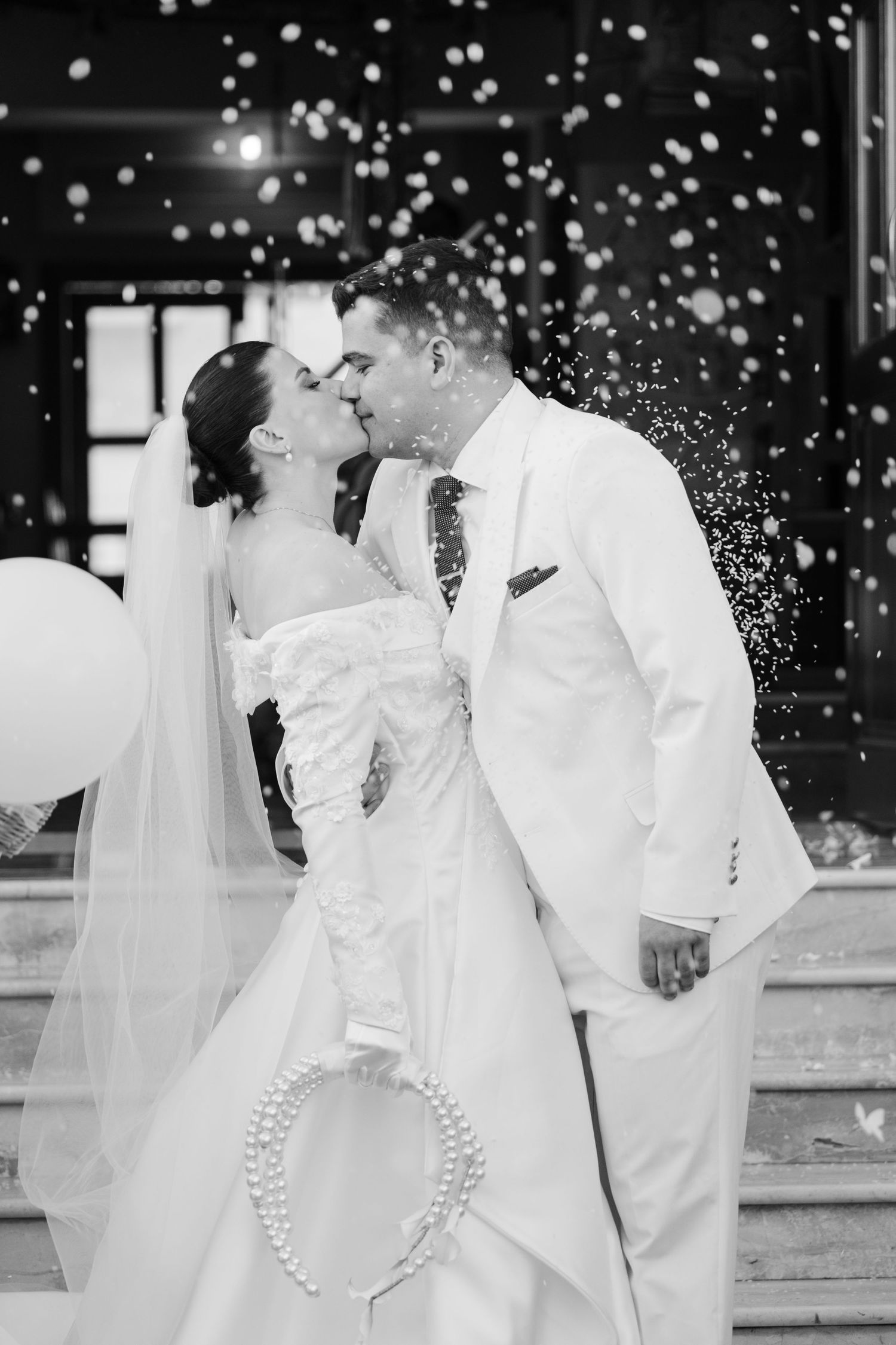 Newlyweds sharing first kiss on church steps surrounded by falling confetti, black and white.