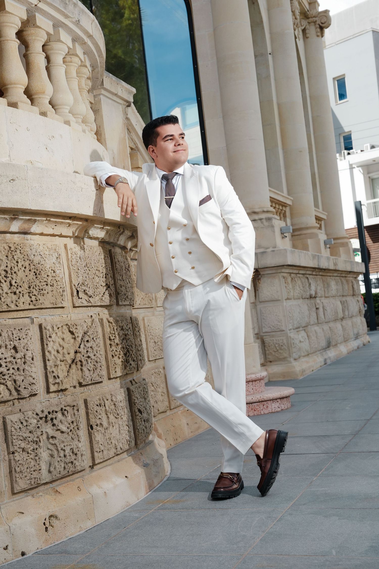 Stylish groom in a white three-piece suit leaning against a stone column of a classical building.