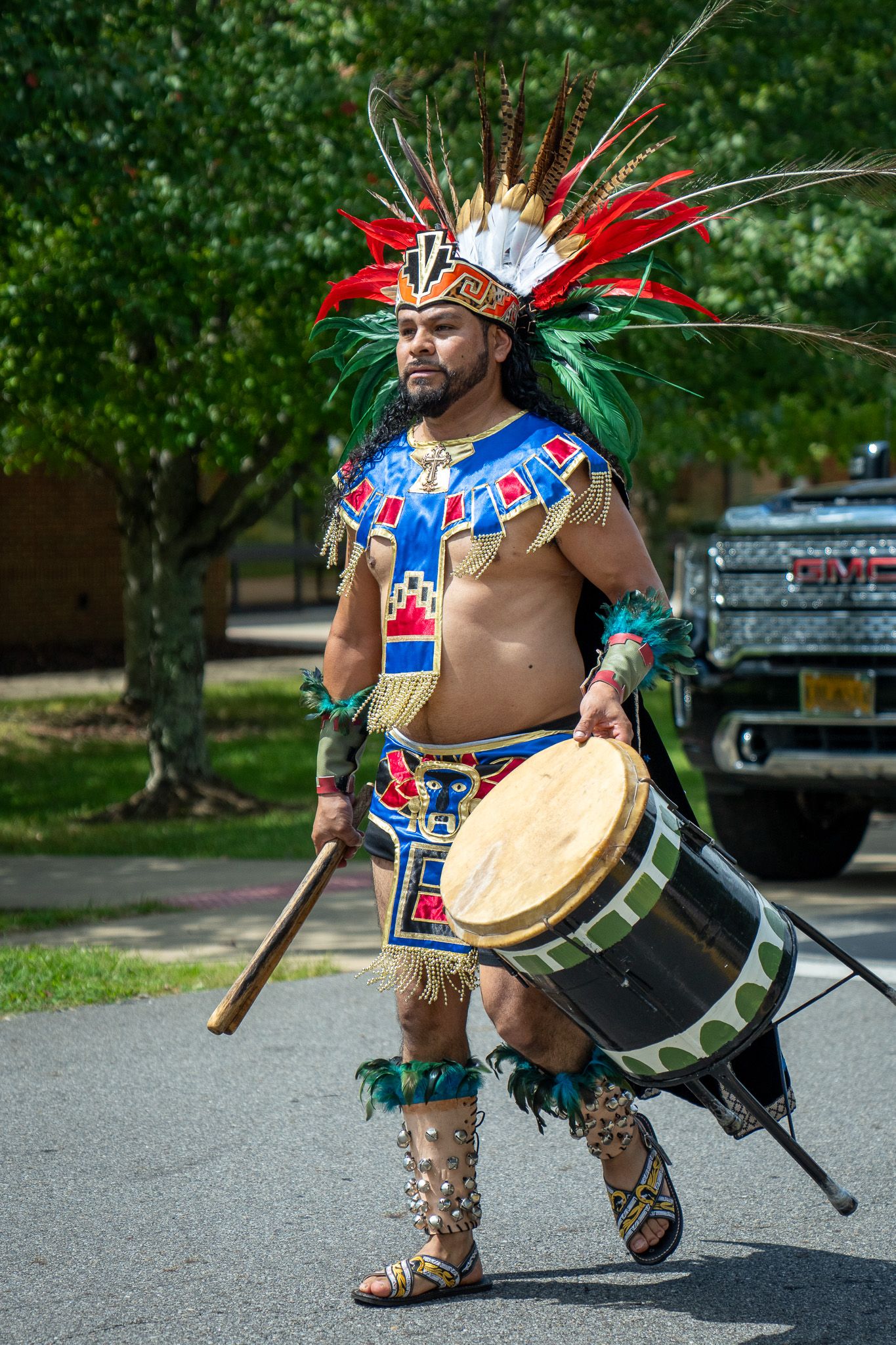 Traditional Native American performer in ceremonial regalia with feathered headdress and drum at outdoor cultural event.